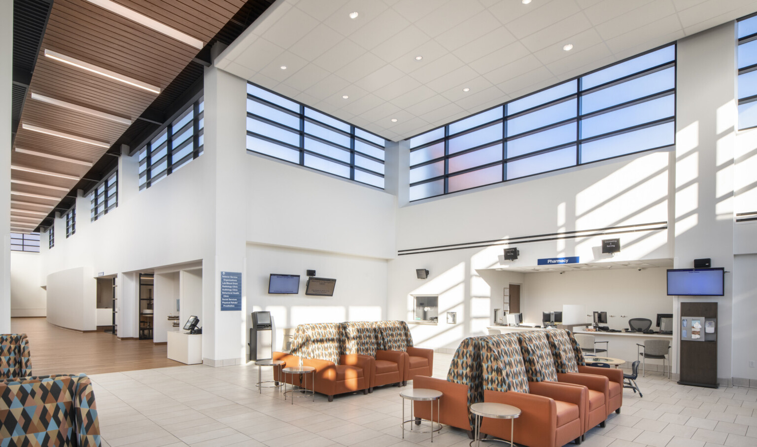 White room with double height ceiling and band of translucent windows around top. Armchair waiting area in front of pharmacy