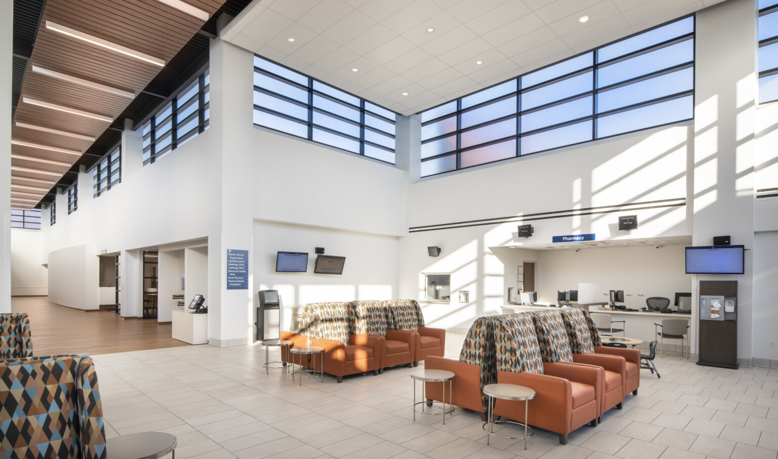 White room with double height ceiling and band of translucent windows around top. Armchair waiting area in front of pharmacy