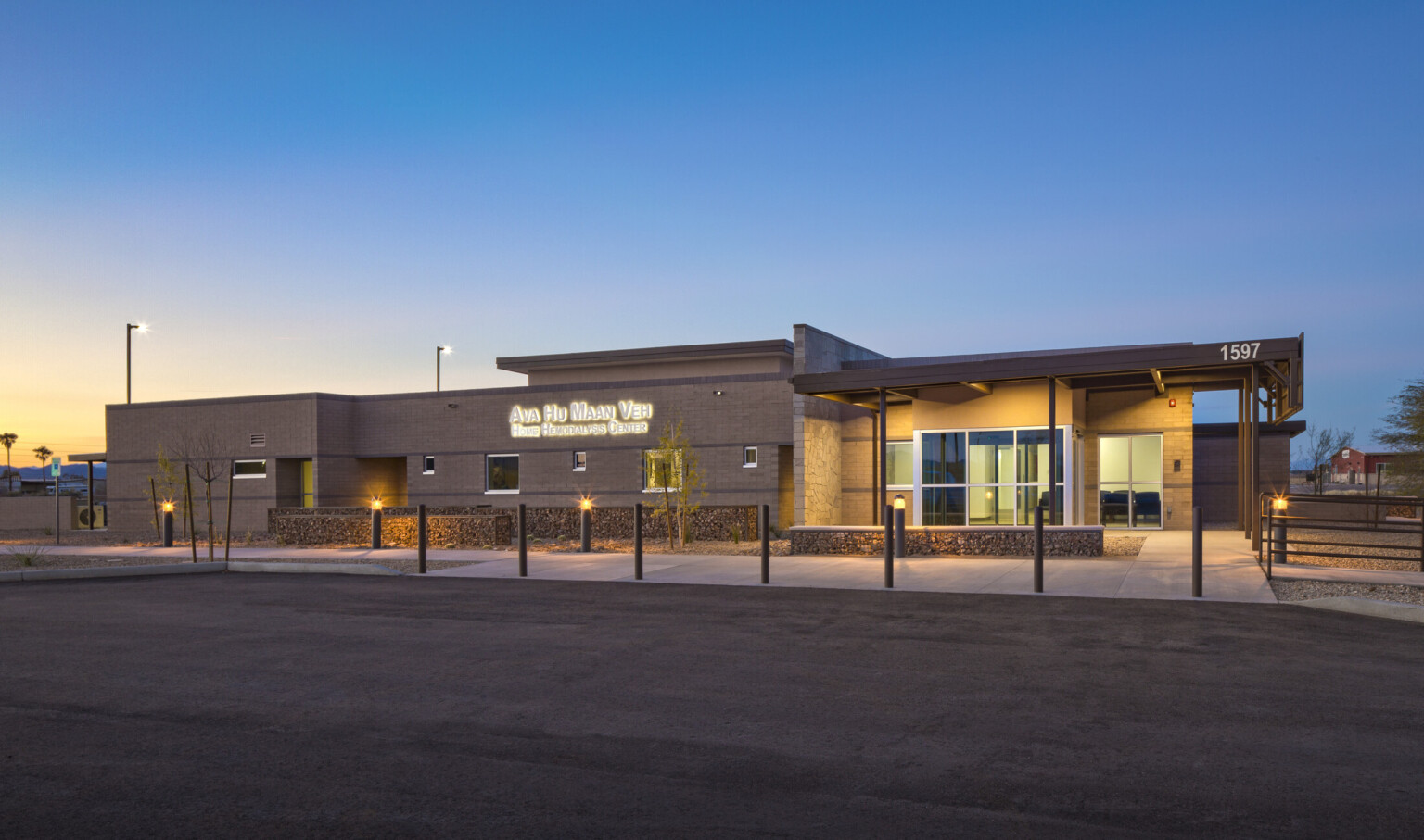 Front of building illuminated from recessed lights in overhang. Lit sign left reads Ava Hu Maan Veh Home Hemodialysis Center