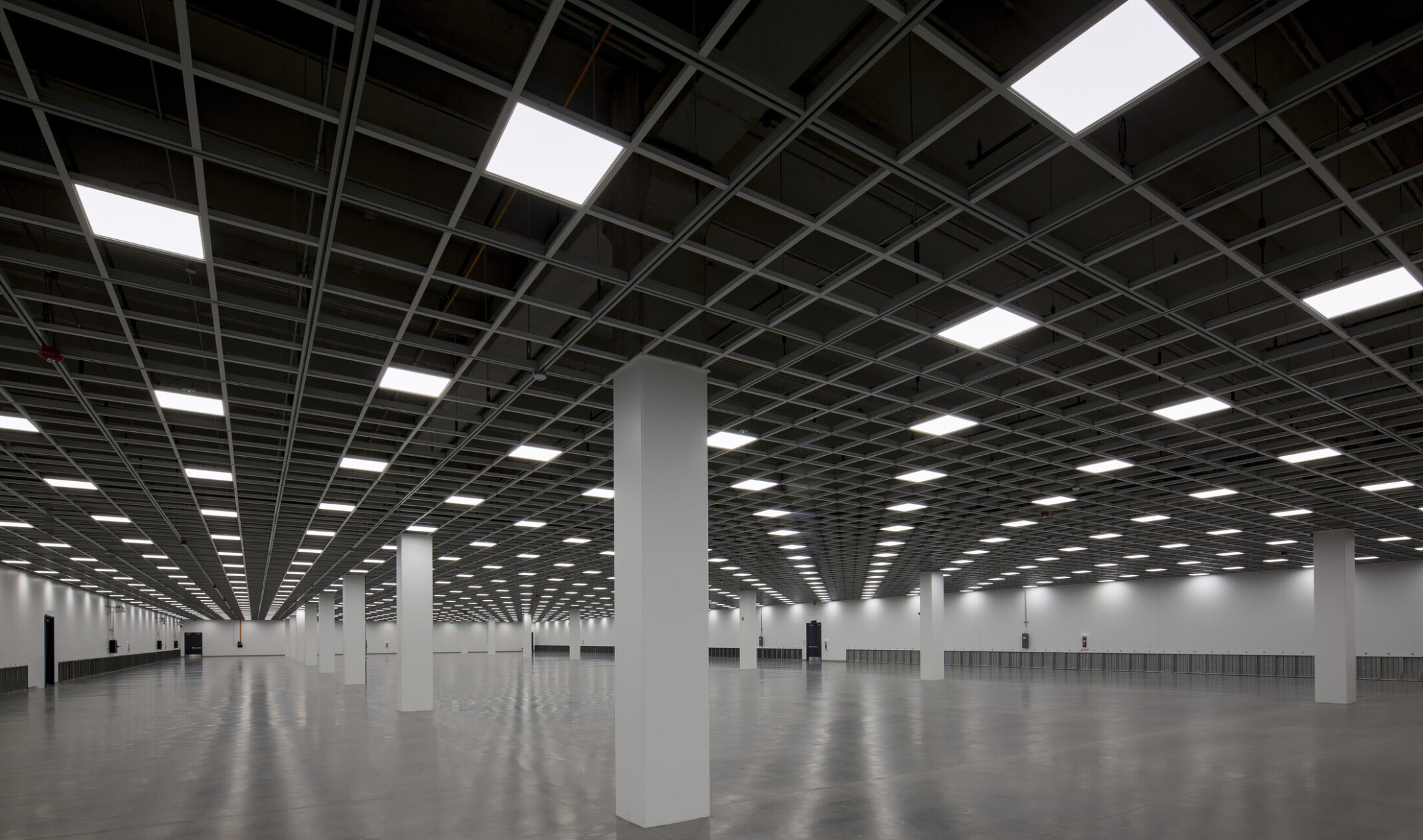 wide open room with reflect cement floors, white square columns white walls, open black ceiling with repeating light squares