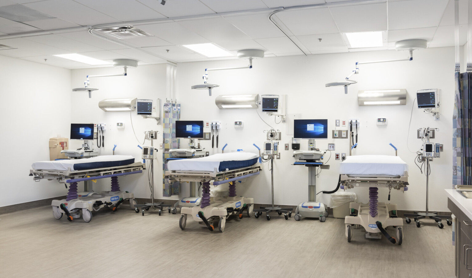 White room with hospital beds, light above, equipment right, and portable computers left. Patterned curtain hangs for privacy
