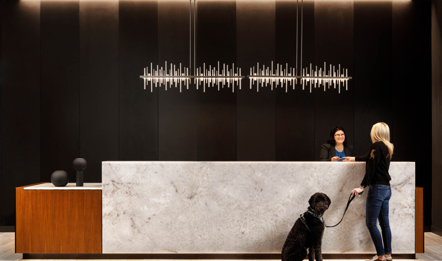 Woman with a black dog at a marble reception desk in front of a black paneled wall. 2 abstract metal lights hang above