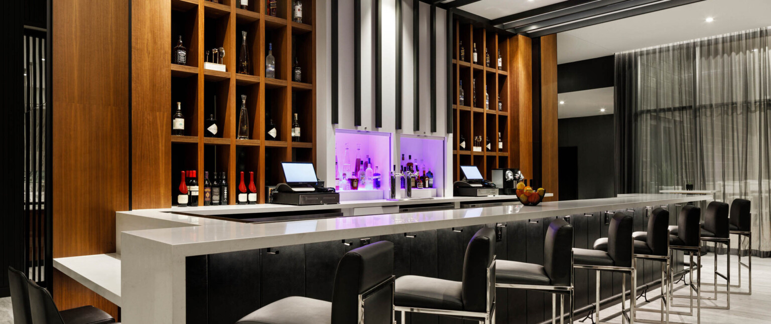Bar with grey counter, black panel base, black bar chairs. Built in shelves behind bar, and white center column lit in purple