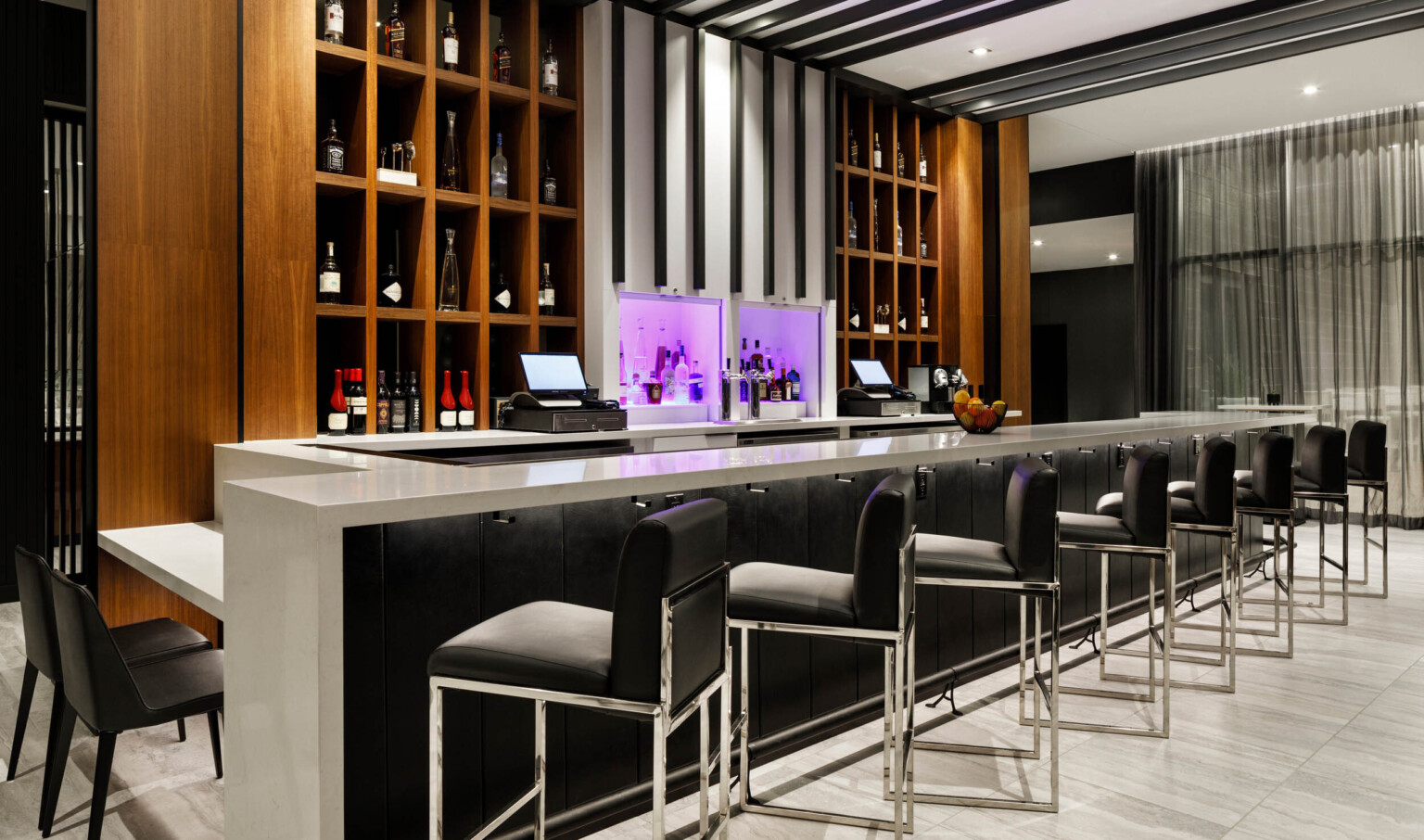 Bar with grey counter, black panel base, black bar chairs. Built in shelves behind bar, and white center column lit in purple