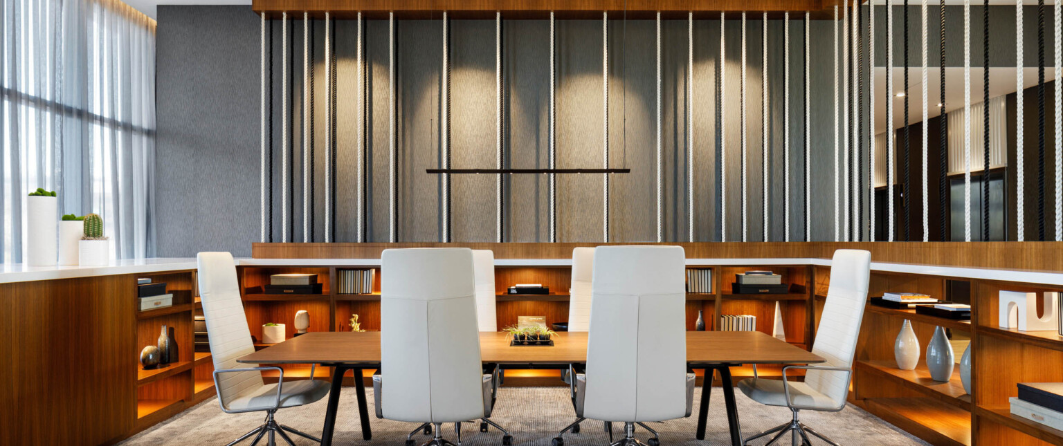 Long wood table with thin white executive chairs in room with wood built in lower shelves with white counter and rope details