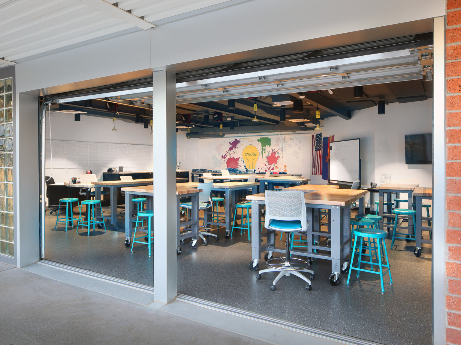 view from exterior: garage door in classroom when retracted brings the outdoors inside, turquoise modern steel stools, light wood tables, office chairs, white walls, polished concrete flooring