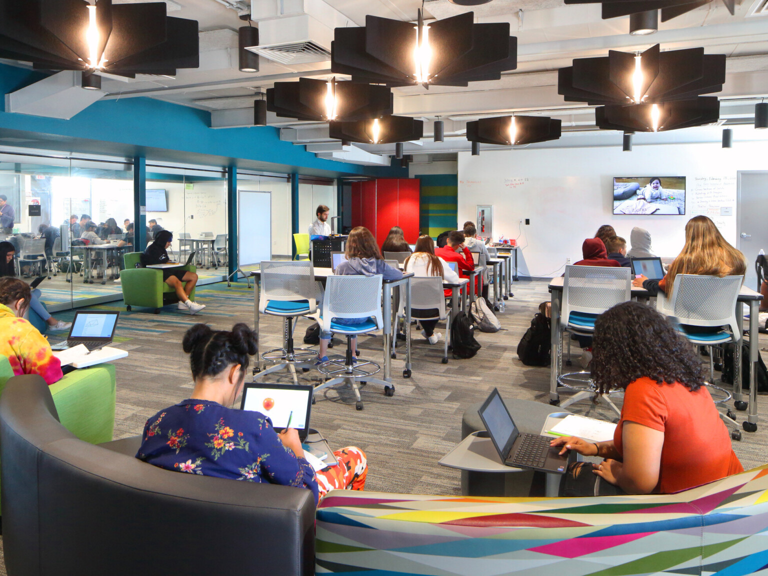 classroom with students in various seating areas, media screen at front, teacher at lectern, overhead edison pendant lights, geometric carpeting, glass partition between two classrooms invite collaboration