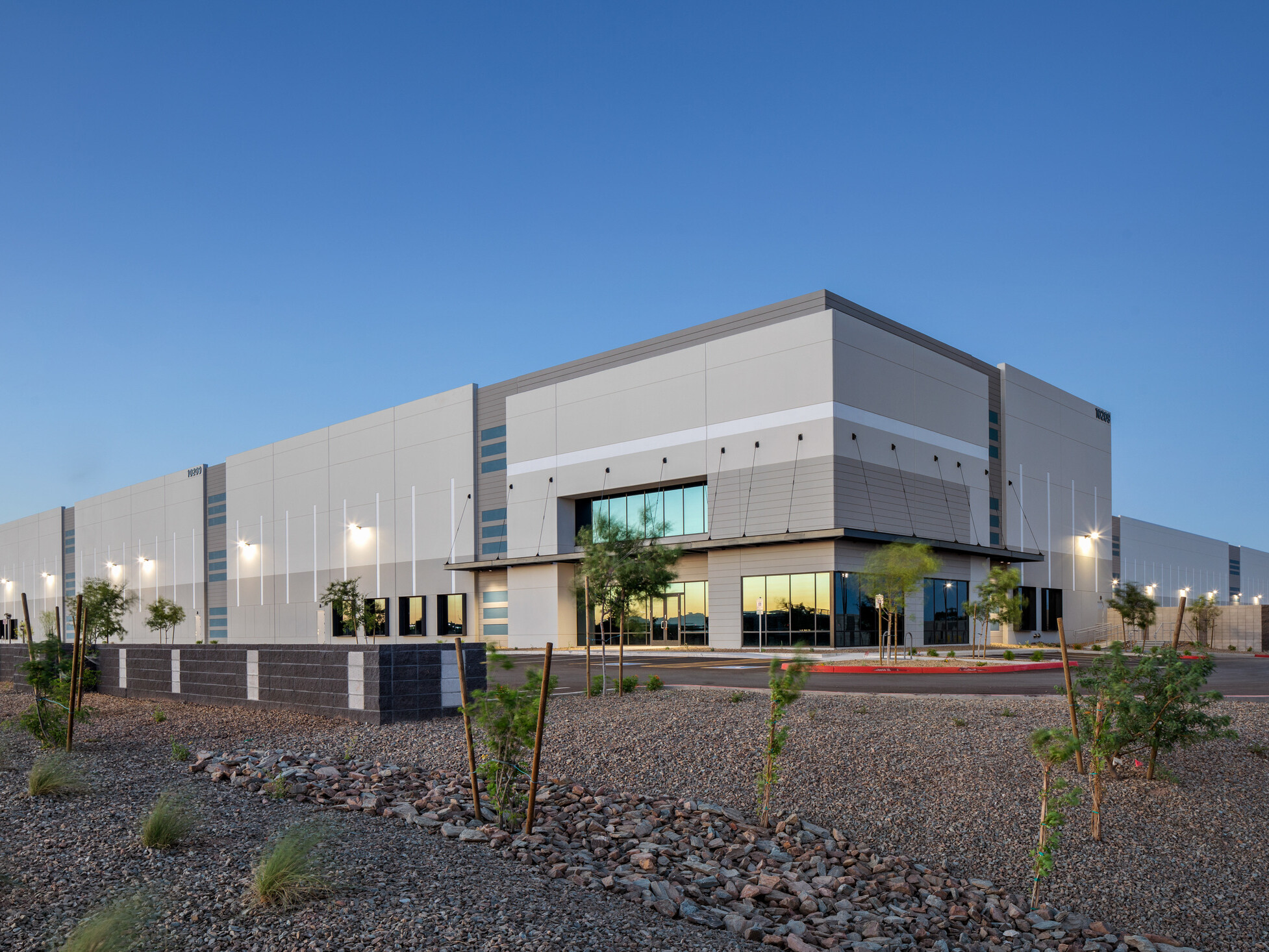 Avondale Commerce Park building exterior; corner view of recessed tenant entry with slatted metal canopy; light to medium gray tilt-up wall panels with blue accent stripe groupings; punched windows marking potential office area; desert landscaping with rock groundcover and sparse plantings