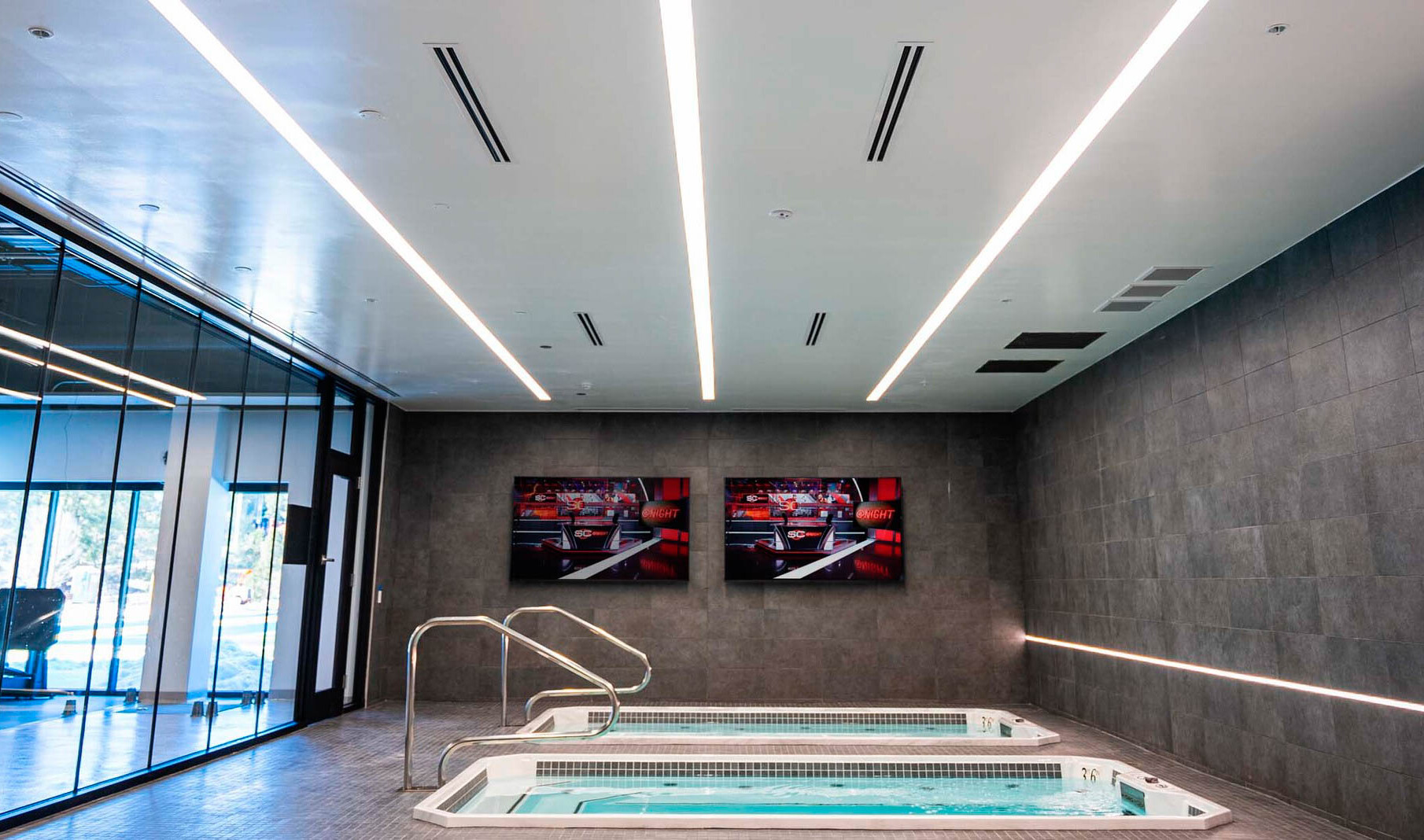 two hydrotherapy pools side by side in a room with white ceiling, stone tiled wall with lighting in it and glass wall.