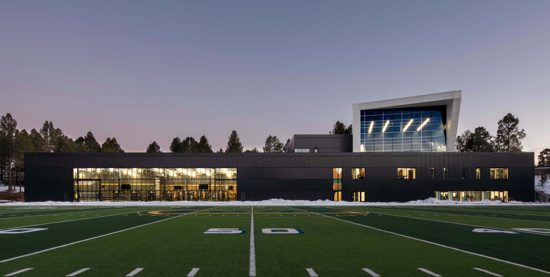 football field in front of a single story building with a multi story angular volume behind it against tree line sky at dusk at Northern Arizona University