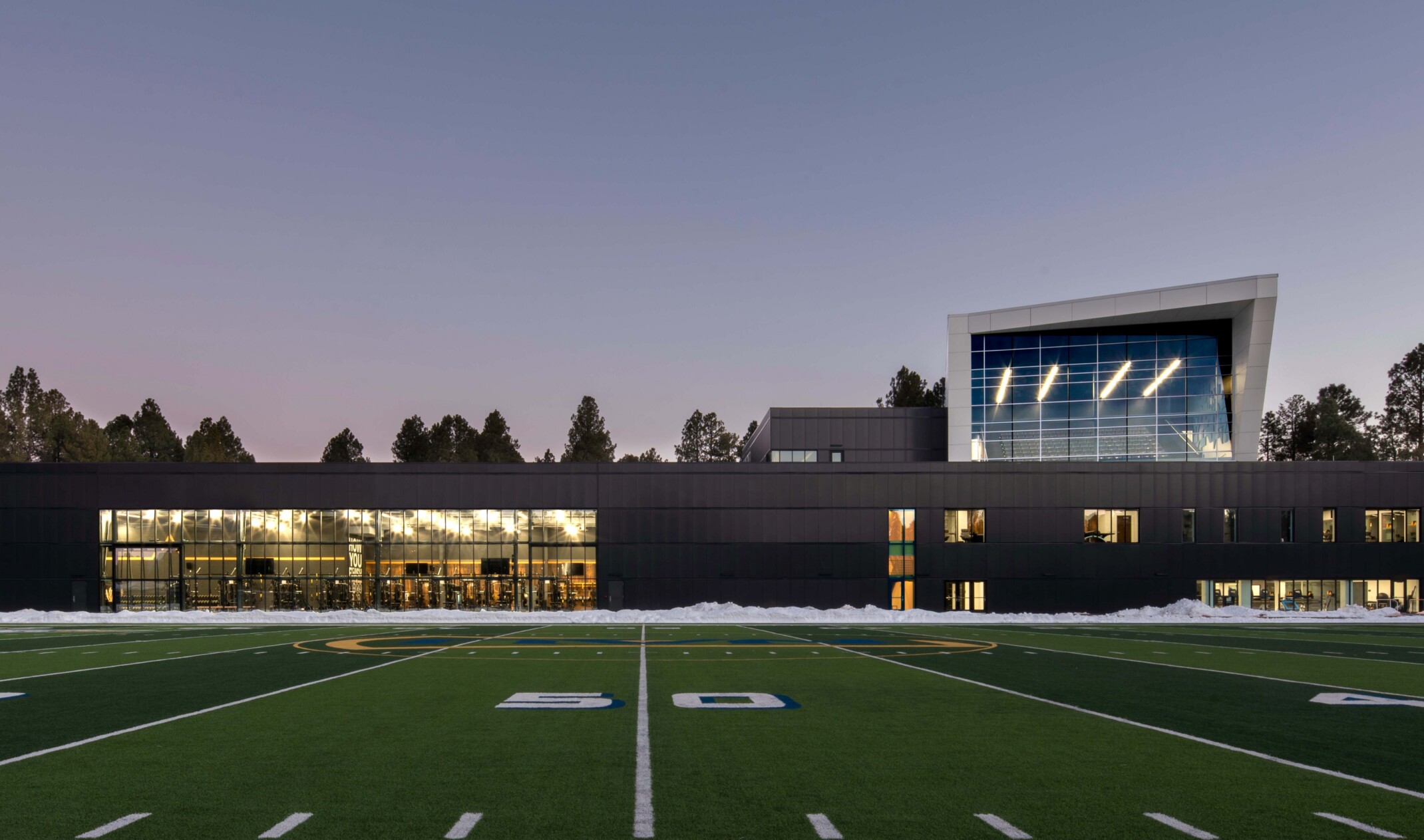 football field in front of a single story building with a multi story angular volume behind it against tree line sky at dusk at Northern Arizona University