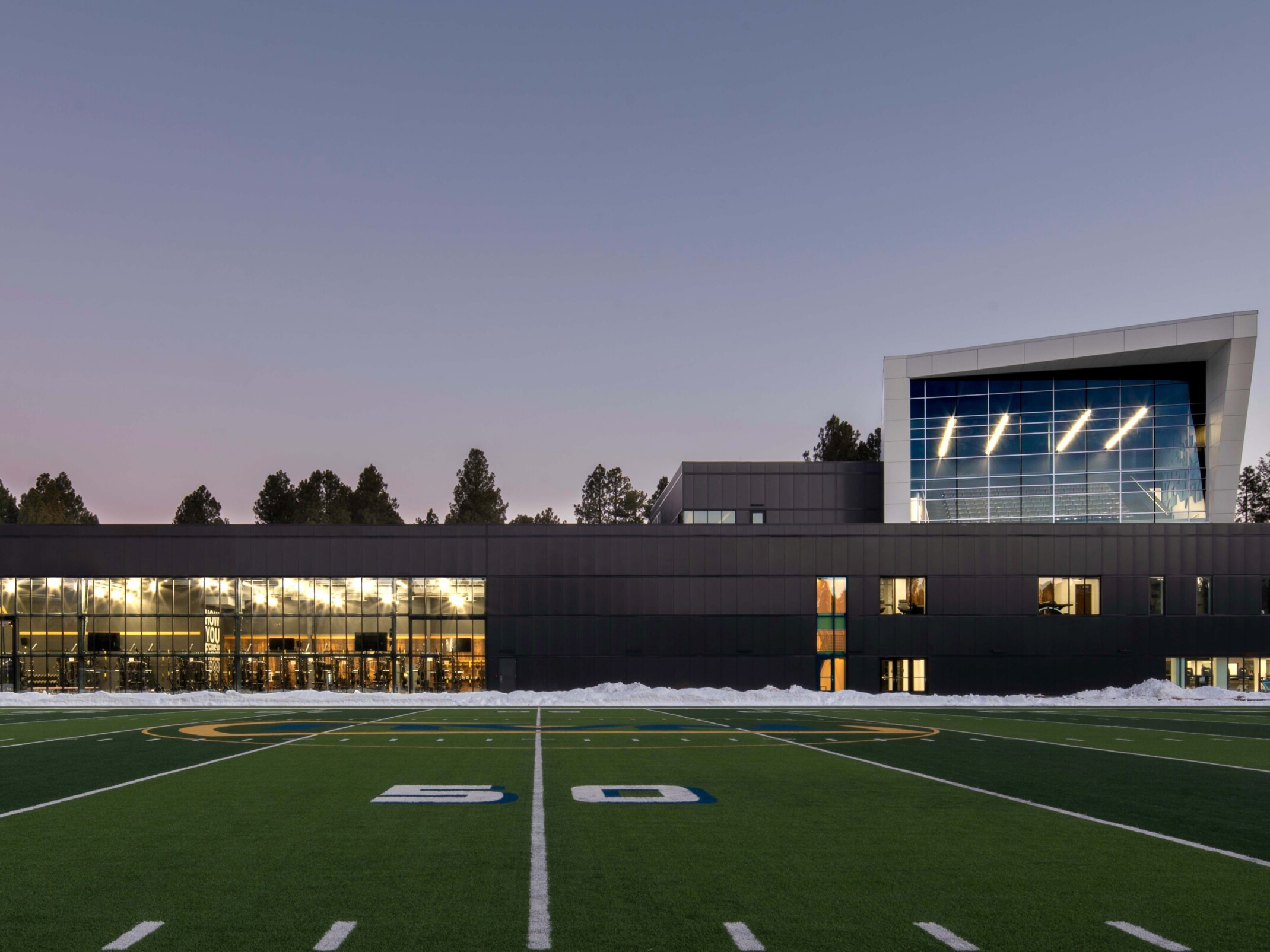 football field in front of a single story building with a multi story angular volume behind it against tree line sky at dusk at Northern Arizona University