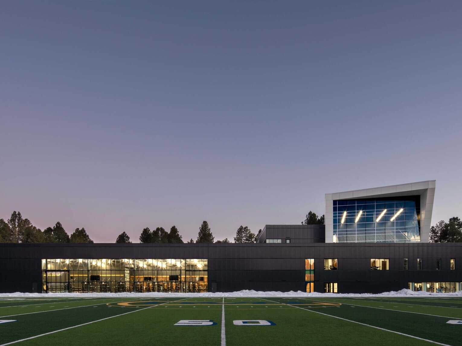 football field in front of a single story building with a multi story angular volume behind it against tree line sky at dusk at Northern Arizona University