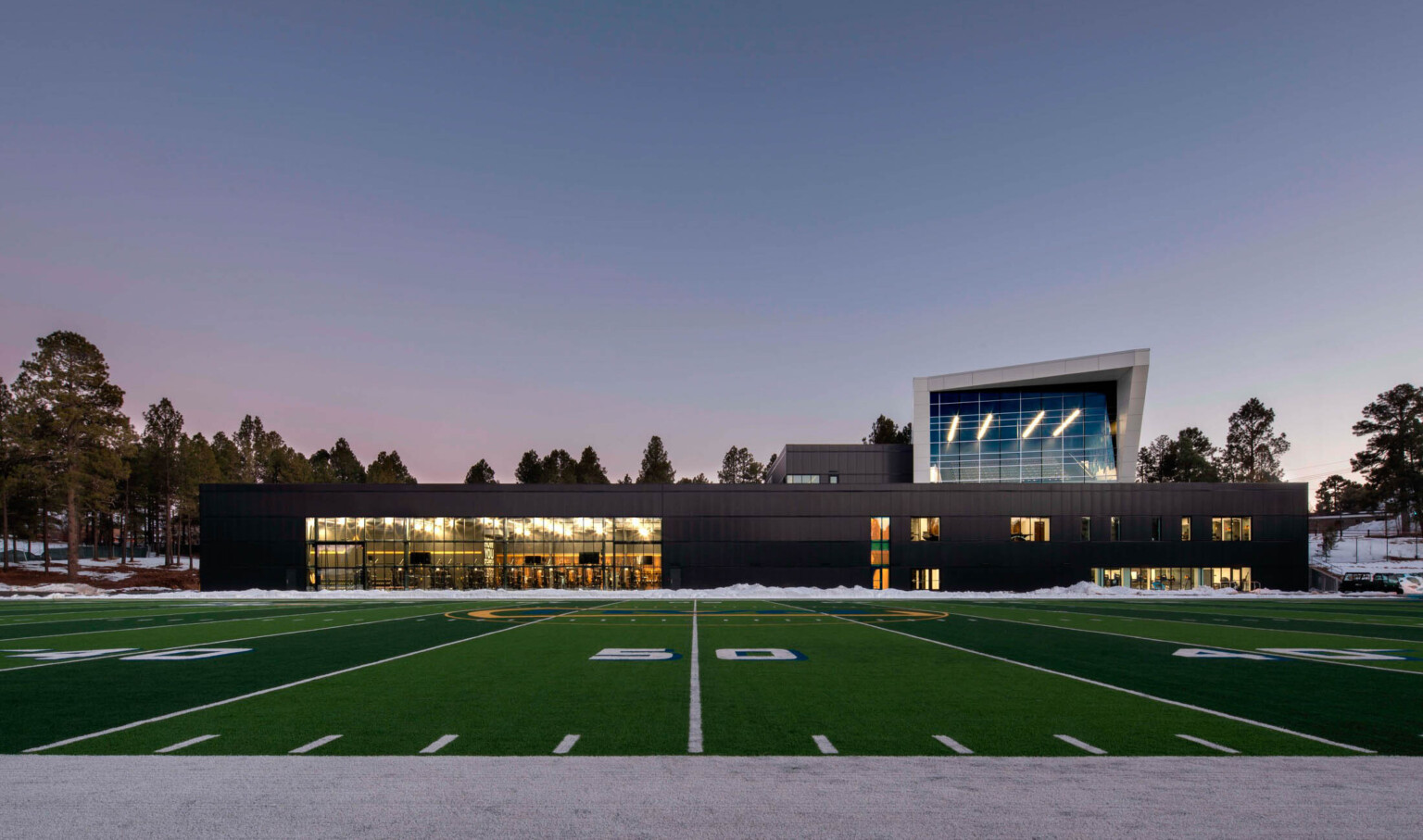 football field in front of a single story building with a multi story angular volume behind it against tree line sky at dusk at Northern Arizona University