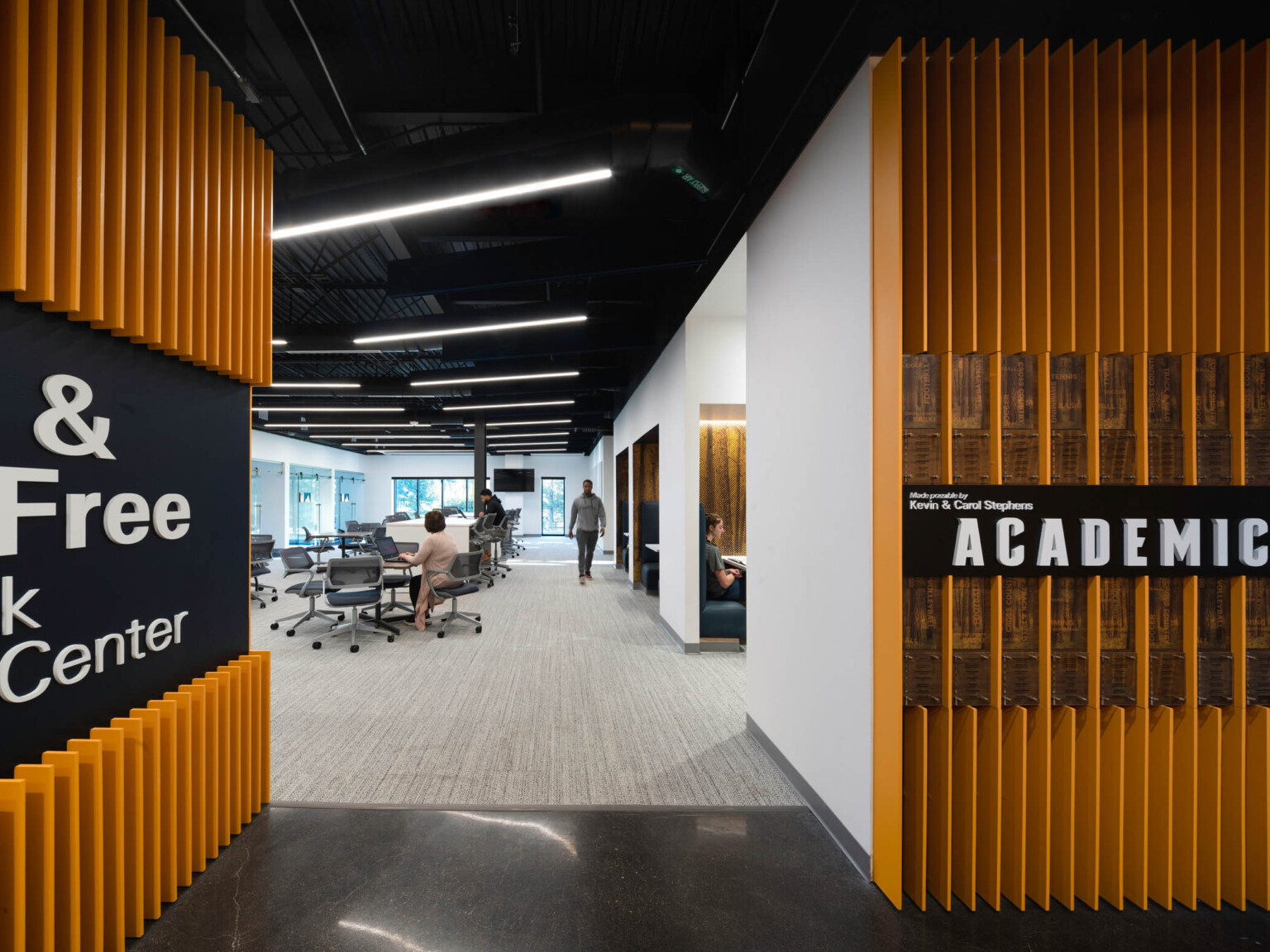 walls covered in yellow vertical fins and white lettering lead to open room with white lights black ceiling and work tables