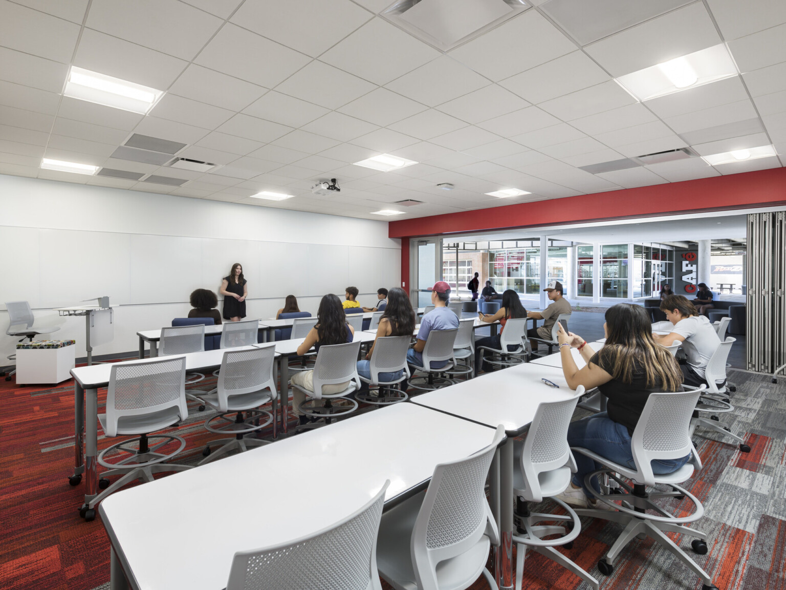 classroom with students in various seating areas, instructor, overhead edison pendant lights, red geometric carpeting, red accent wall, glass partition between two classrooms invite collaboration