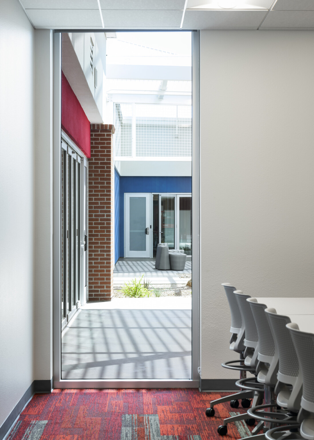 view from classroom to outside quad through floor-to-ceiling window, red geometric carpeting, white tables and desk chairs