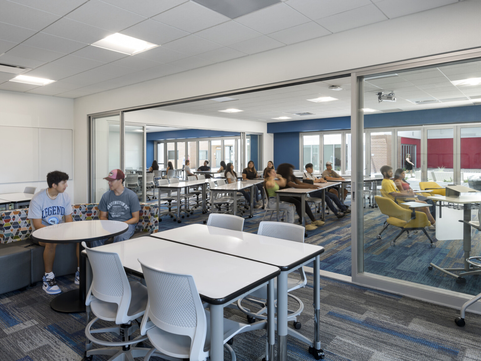 study area outside classroom with students in various seating areas, instructor, overhead lighting, blue and grey geometric carpeting, blue accent wall, glass partition between two classrooms invite collaboration