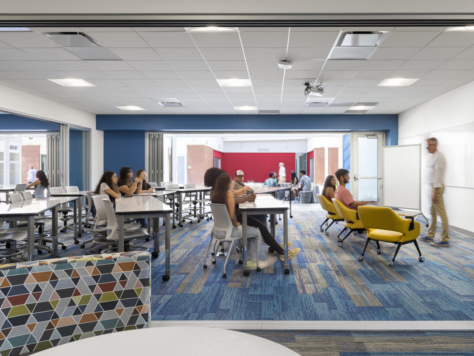 classroom with students in various seating areas, instructor, overhead edison pendant lights, blue geometric carpeting, blue accent wall, glass partition between two classrooms invite collaboration
