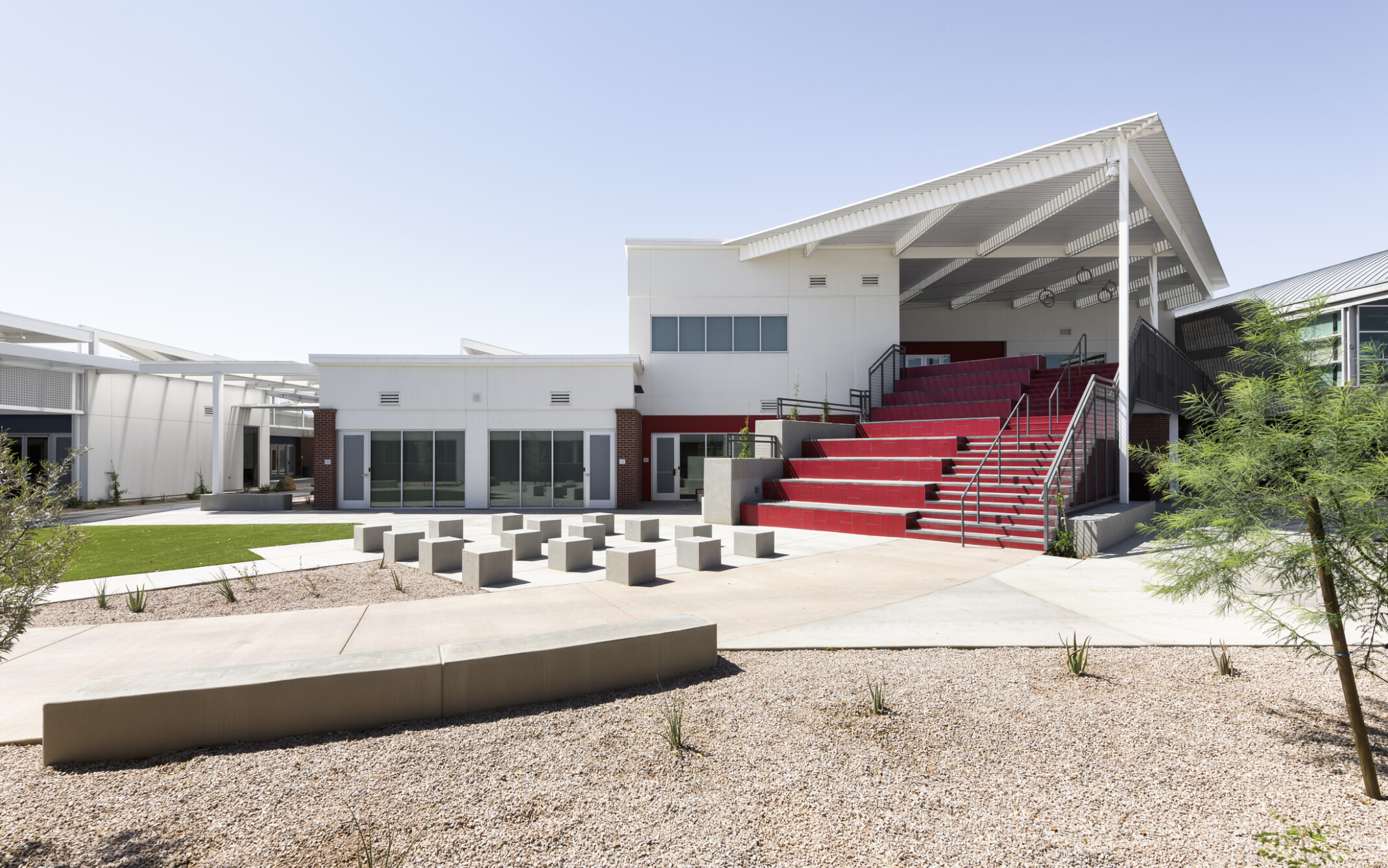 exterior of the quad and connected buildings, split section red stairs with steps and seating, white building with angular steel roof, concrete pathways, pea gravel landscaping, trees, grass