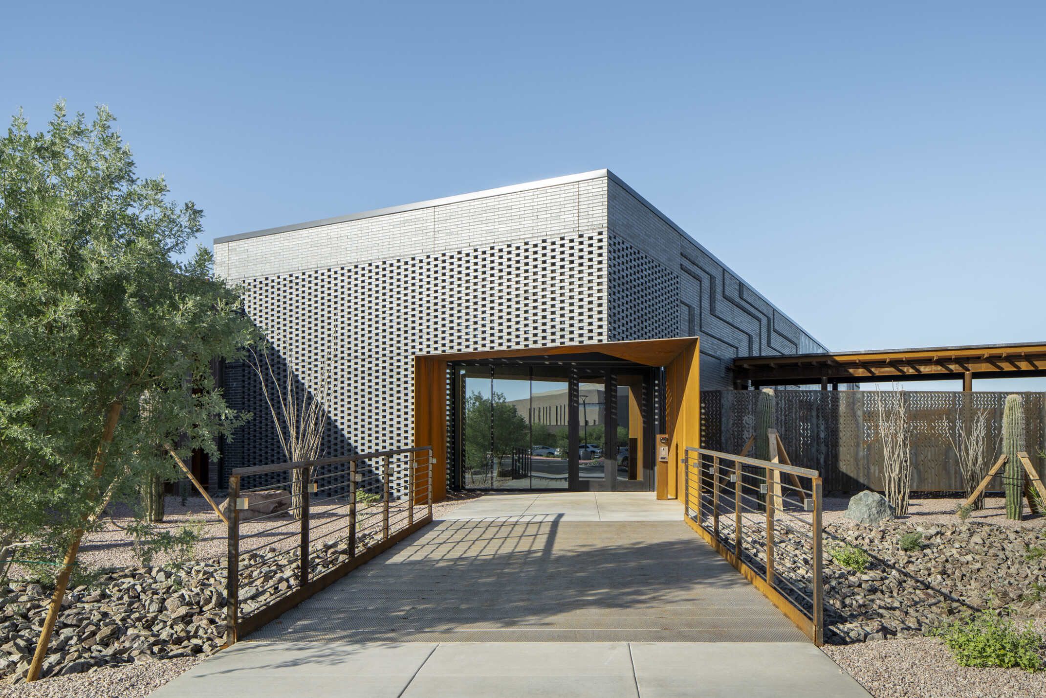 Entrance to a building with a textured gray brick and patterned perforated panels, adding depth and visual interest; Bright daylight casts sharp shadows across the walkway, emphasizing the linear design elements
