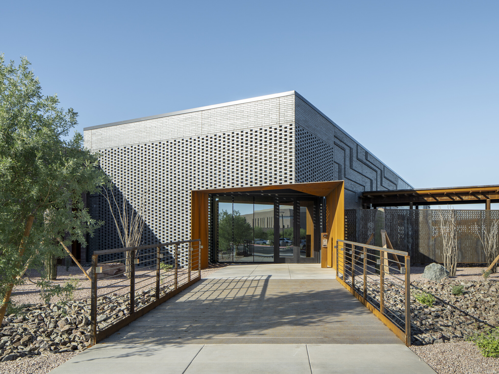 Entrance to a building with a textured gray brick and patterned perforated panels, adding depth and visual interest; Bright daylight casts sharp shadows across the walkway, emphasizing the linear design elements
