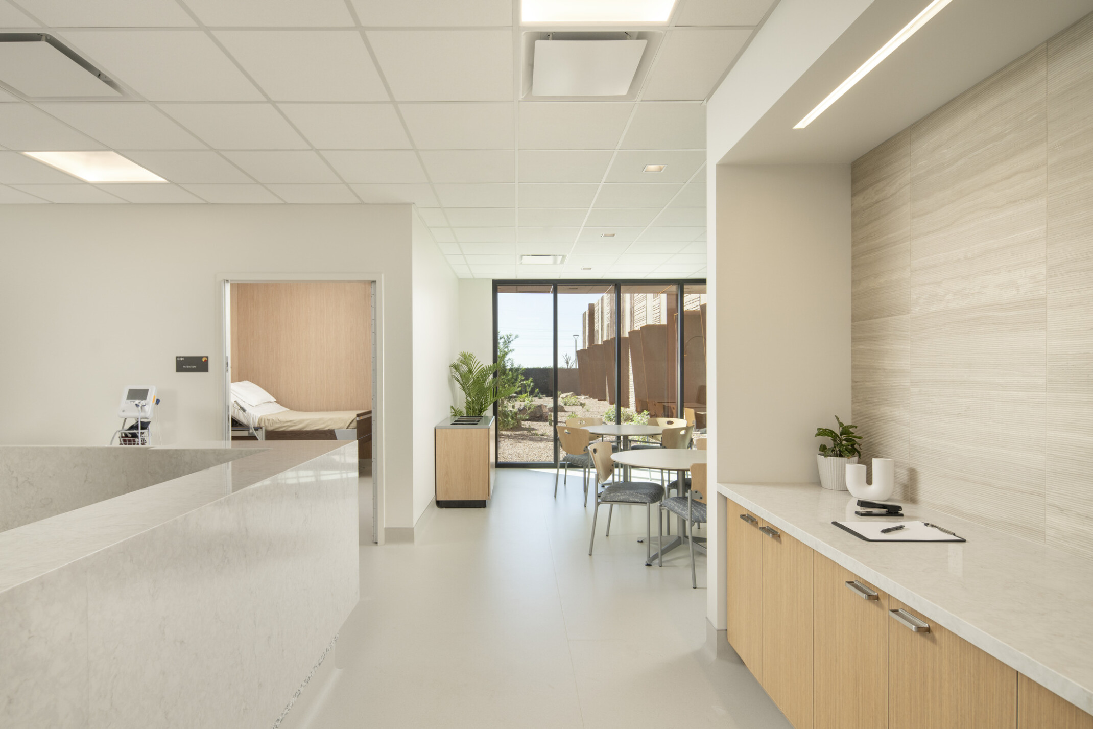 A bright, modern interior space with a clean, minimalist design; white marble reception counter on the left and a light wood cabinet with a white countertop on the right, decorated with small potted plants and a clipboard; grid-patterned ceiling and recessed lighting; desert landscaping and rust-colored metal panels. A small round table with chairs is positioned near the glass door, and through an open doorway on the left, a hospital bed is partially visible in a private room