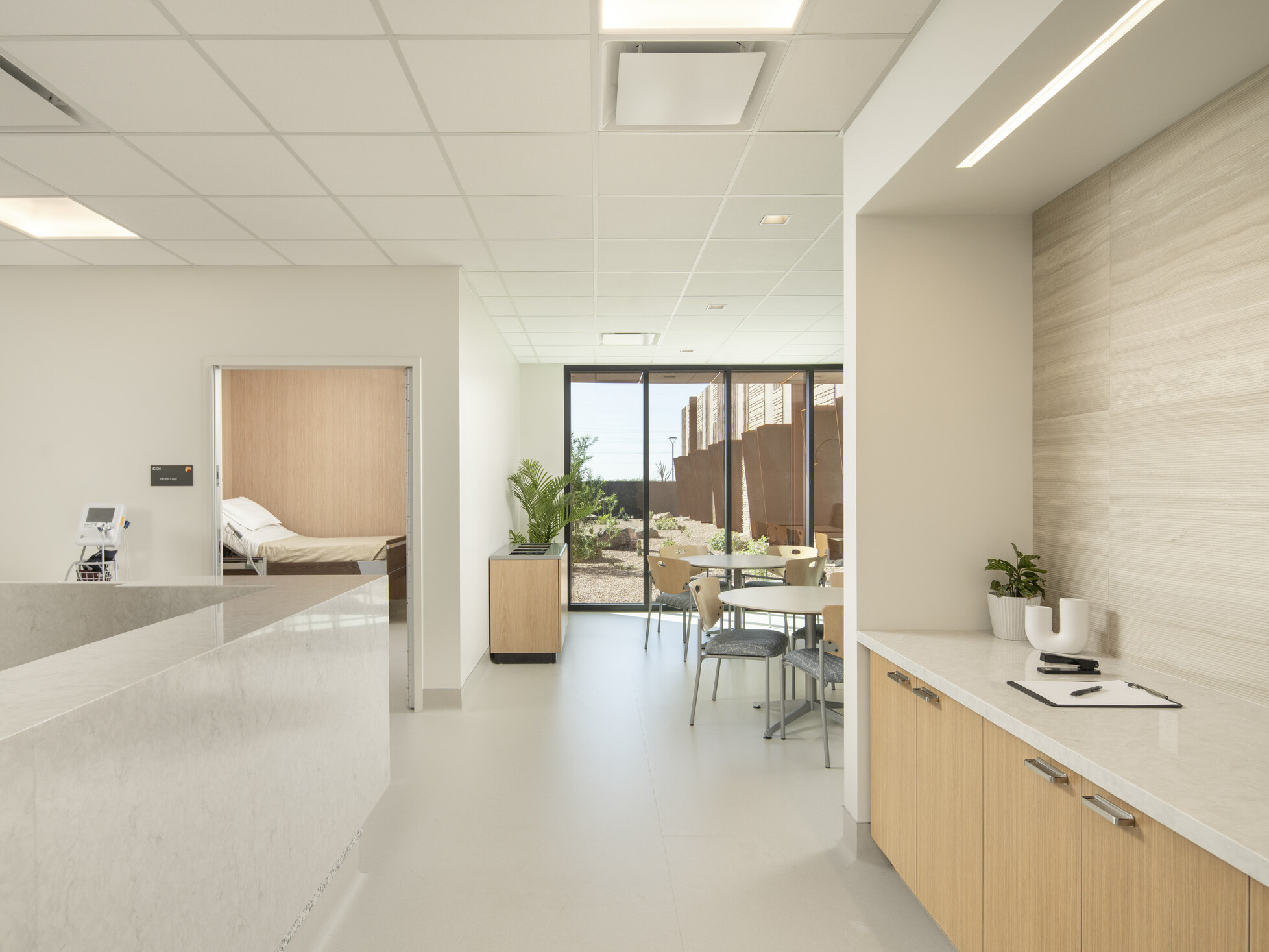 A bright, modern interior space with a clean, minimalist design; white marble reception counter on the left and a light wood cabinet with a white countertop on the right, decorated with small potted plants and a clipboard; grid-patterned ceiling and recessed lighting; desert landscaping and rust-colored metal panels. A small round table with chairs is positioned near the glass door, and through an open doorway on the left, a hospital bed is partially visible in a private room