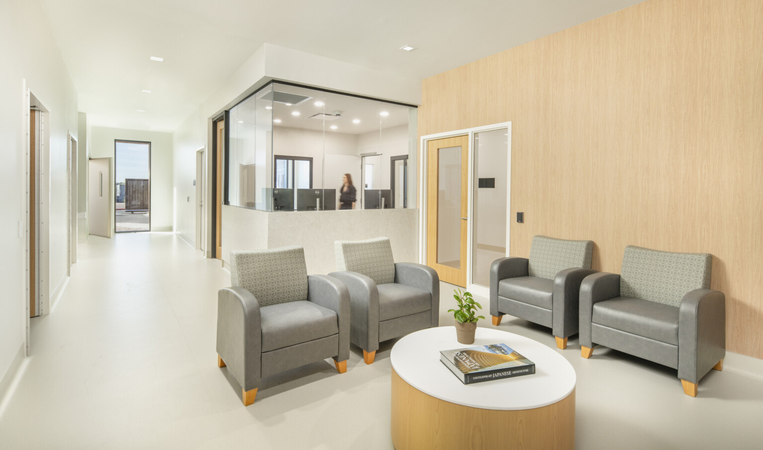 A bright lobby area with four upholstered chairs arranged around a round table, adjacent to a glass-walled office space and a hallway extending toward an exterior doorway.