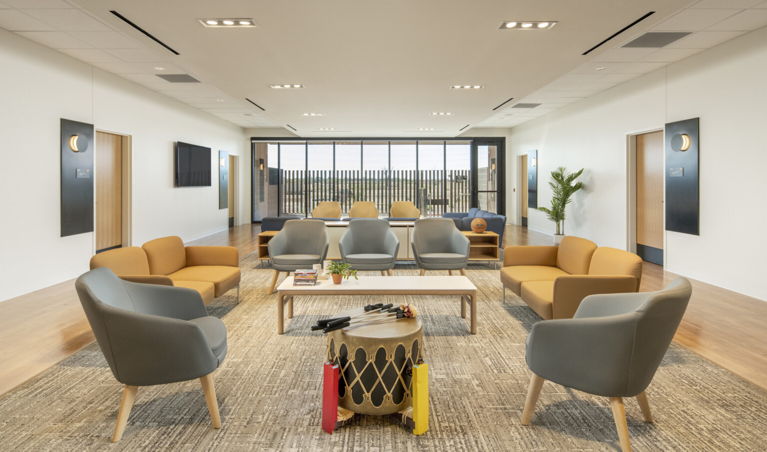 A spacious lounge with gray and yellow seating arranged around low tables, a drum in the center, wood flooring, and large windows at the far end letting in natural light.