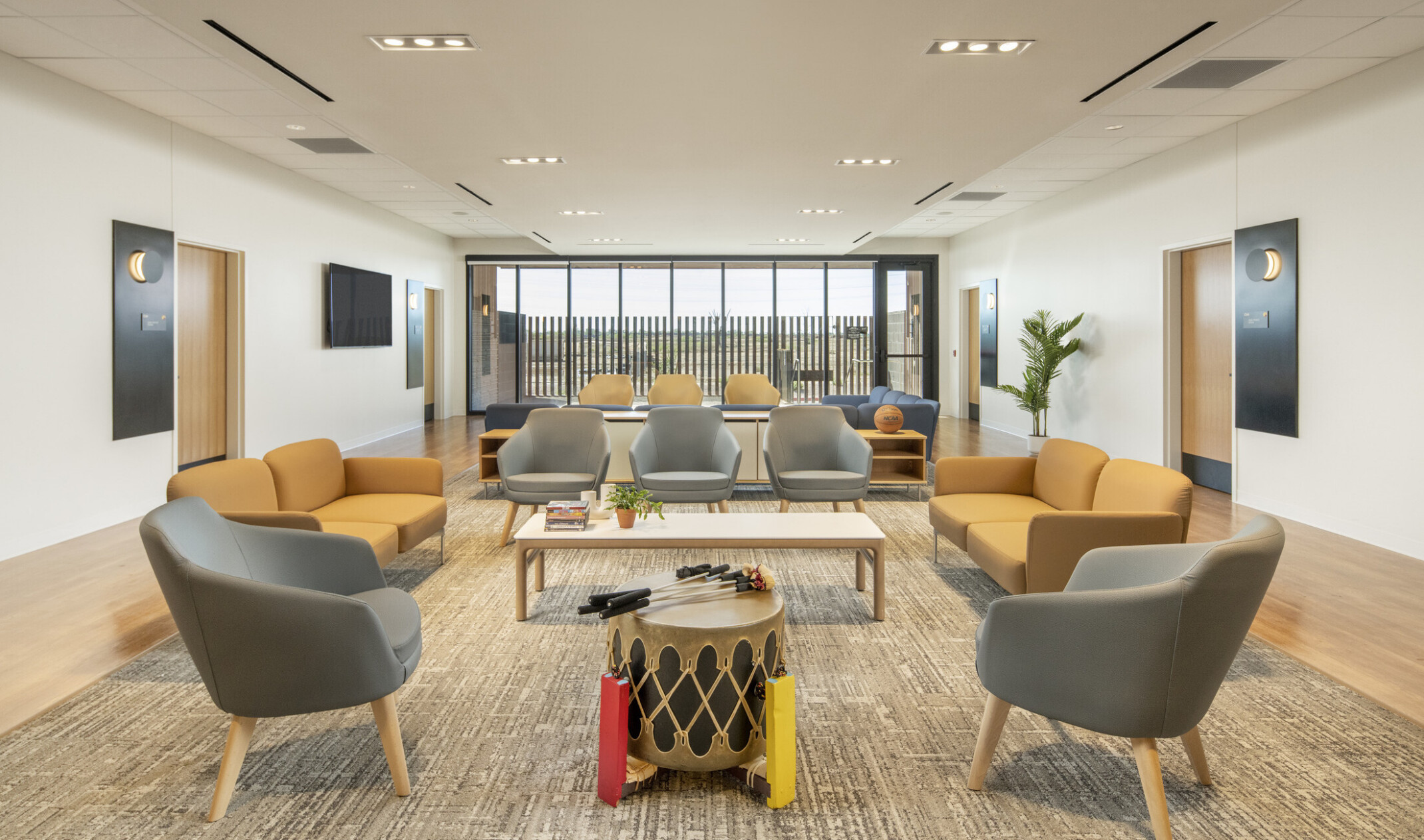 A spacious lounge with gray and yellow seating arranged around low tables, a drum in the center, wood flooring, and large windows at the far end letting in natural light.