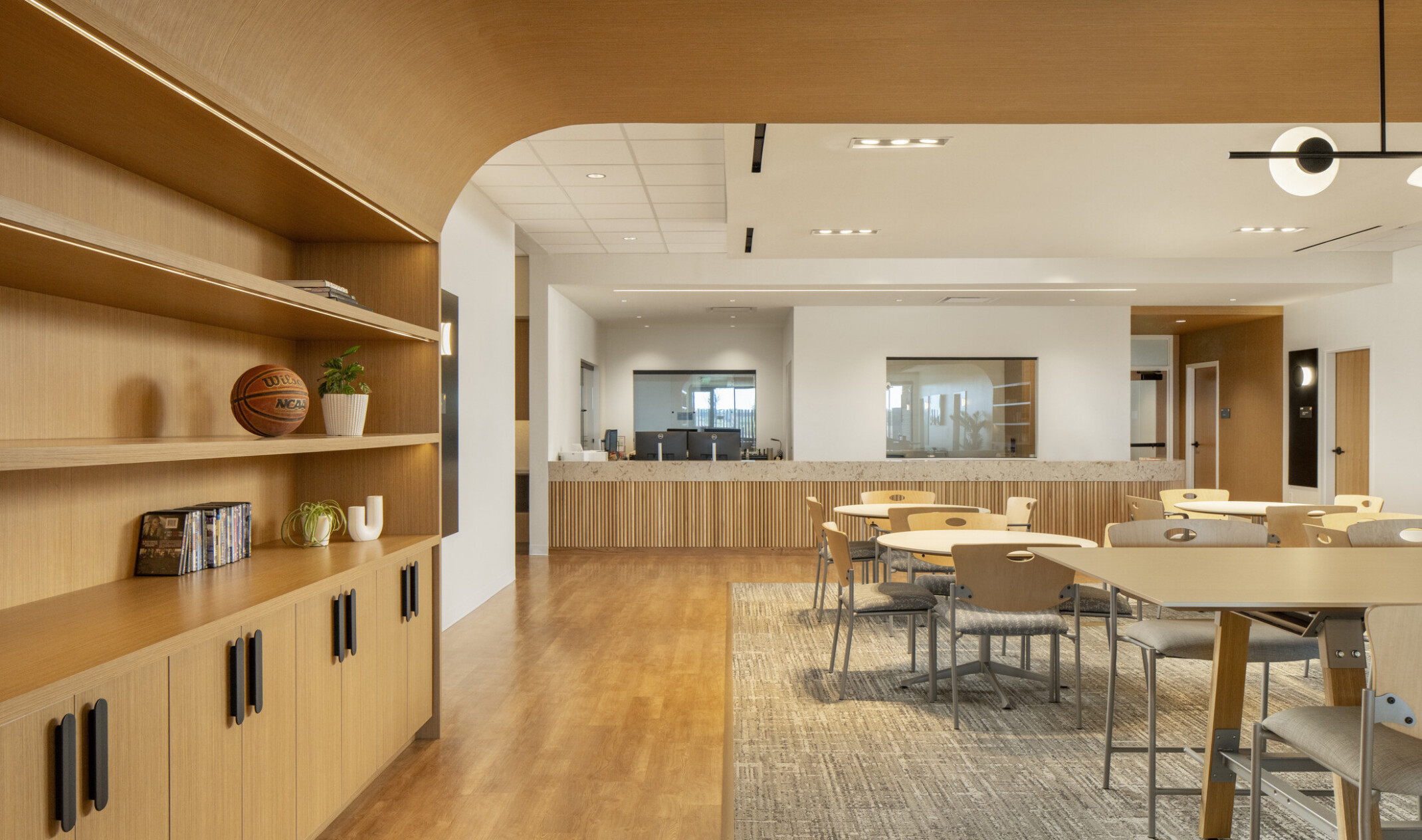 Modern open-concept office lounge with light wood finishes, featuring built-in shelving with decorative items on the left and multiple round tables with chairs on a large area rug. The space has wood flooring, white walls, and a mix of recessed and pendant lighting, creating a warm and contemporary atmosphere.