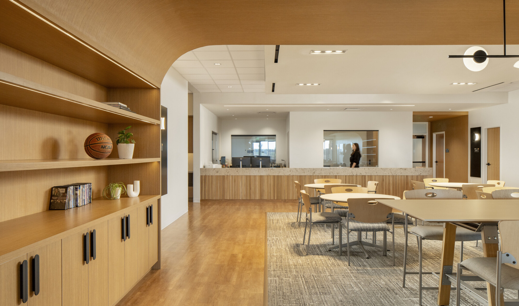 Modern open-concept office lounge with light wood finishes, featuring built-in shelving with decorative items on the left and multiple round tables with chairs on a large area rug. The space has wood flooring, white walls, and a mix of recessed and pendant lighting, creating a warm and contemporary atmosphere.
