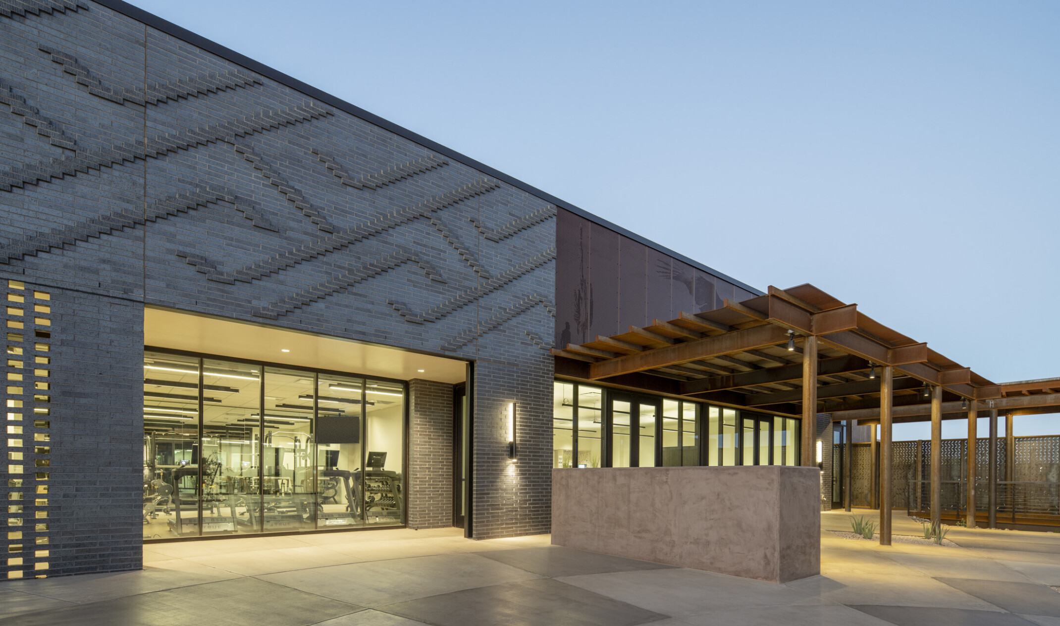 A modern, angular building with a minimalist design; sunset; textured dark gray concrete panels and rust-colored metal accents, creating a striking contrast. Large floor-to-ceiling glass windows line the front, reflecting the outdoor landscape; gravel-covered ground with sparse desert plants; contemporary desert aesthetic; warm interior lighting