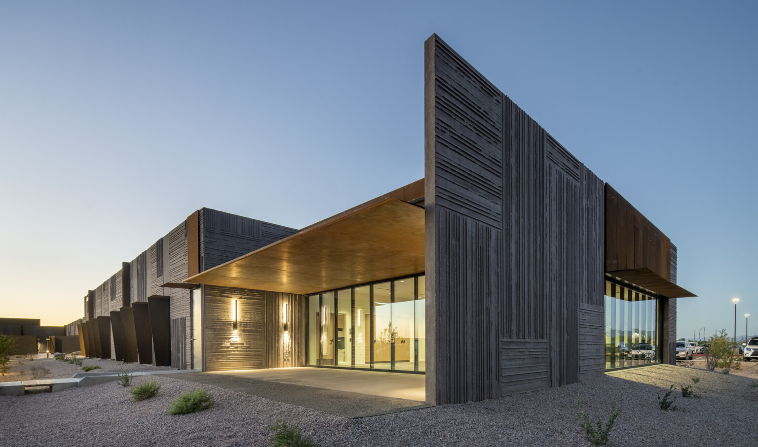 A modern, angular building with a minimalist design; sunset; textured dark gray concrete panels and rust-colored metal accents, creating a striking contrast. Large floor-to-ceiling glass windows line the front, reflecting the outdoor landscape; gravel-covered ground with sparse desert plants; contemporary desert aesthetic; warm interior lighting