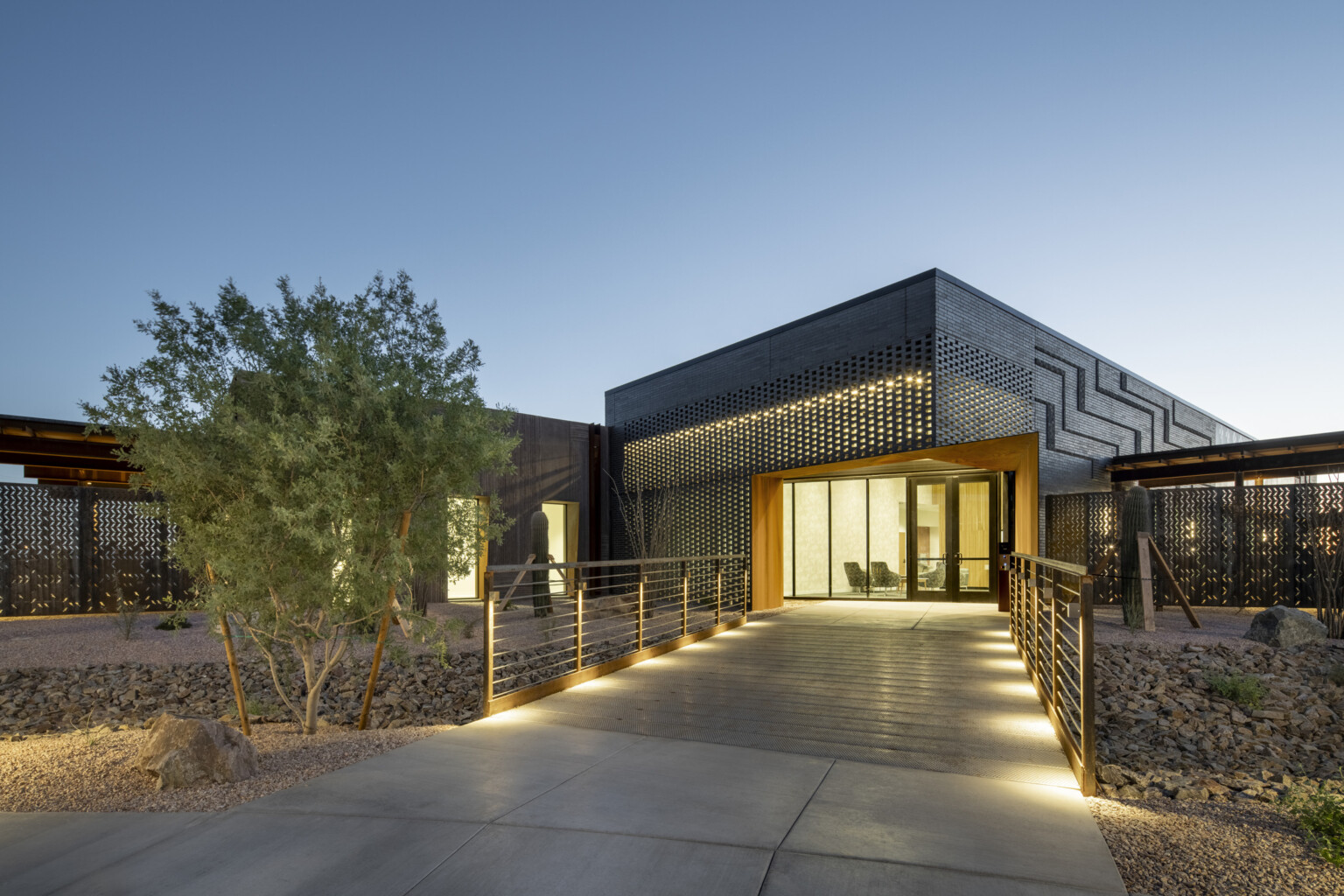 Modern building exterior at dusk with illuminated walkway leading to a glass entrance framed by a rust-colored steel structure; facade features dark perforated metal panels with geometric patterns; desert landscaping with rocks and shrubs surrounds the path under a clear evening sky