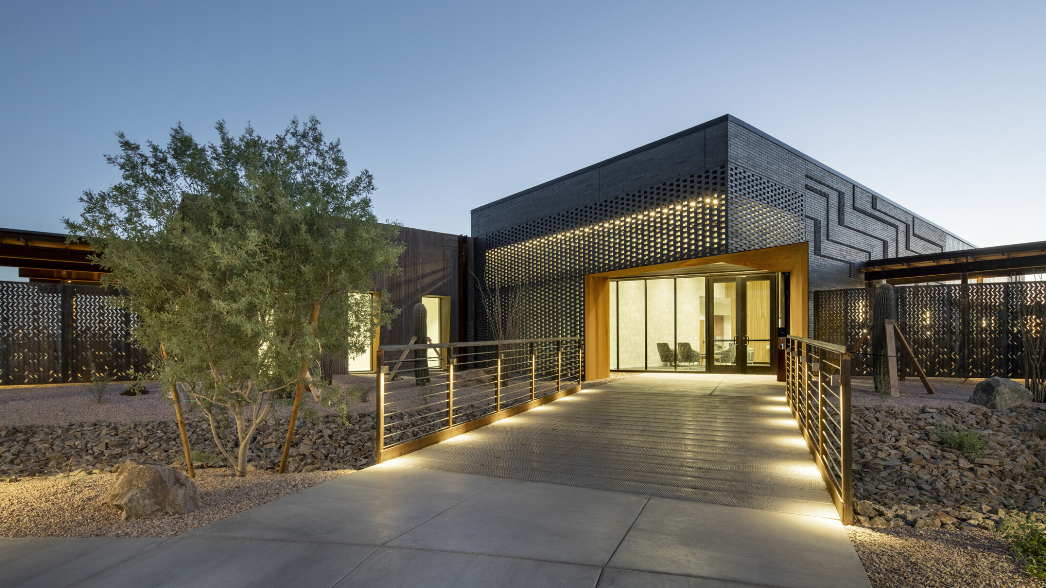 Modern building exterior at dusk with illuminated walkway leading to a glass entrance framed by a rust-colored steel structure; facade features dark perforated metal panels with geometric patterns; desert landscaping with rocks and shrubs surrounds the path under a clear evening sky