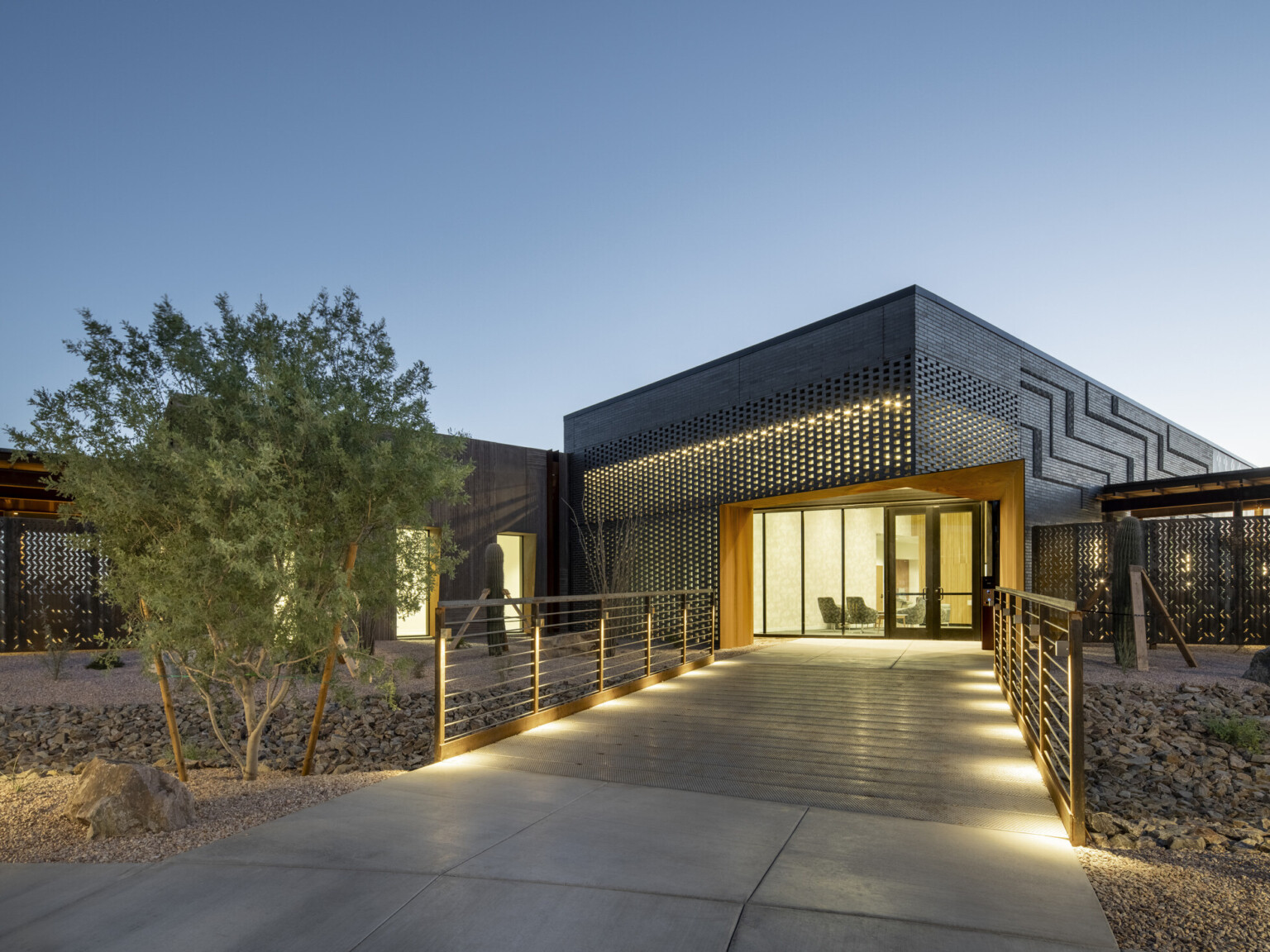 Modern building exterior at dusk with illuminated walkway leading to a glass entrance framed by a rust-colored steel structure; facade features dark perforated metal panels with geometric patterns; desert landscaping with rocks and shrubs surrounds the path under a clear evening sky