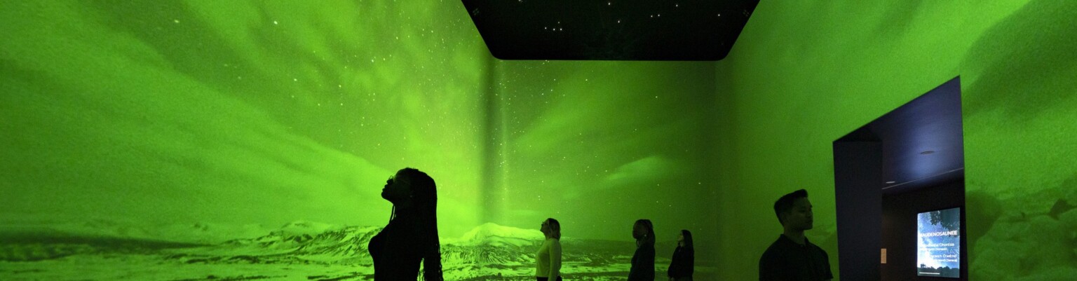 An immersive art exhibit at the Heard Museum with screens on all visible walls of the room showing the Northern Lights among snow-capped hills colorized with bright green