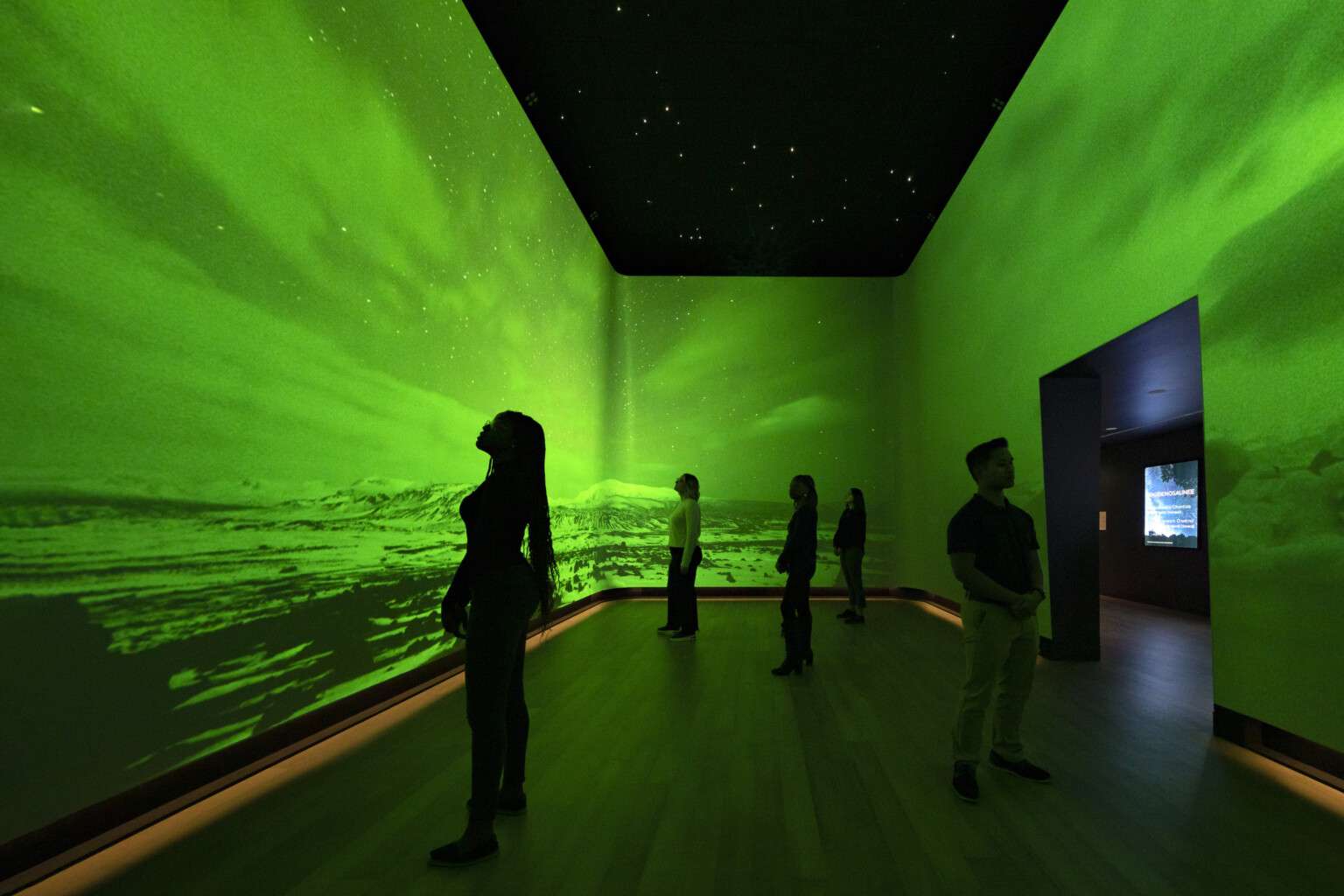 An immersive art exhibit at the Heard Museum with screens on all visible walls of the room showing the Northern Lights among snow-capped hills colorized with bright green