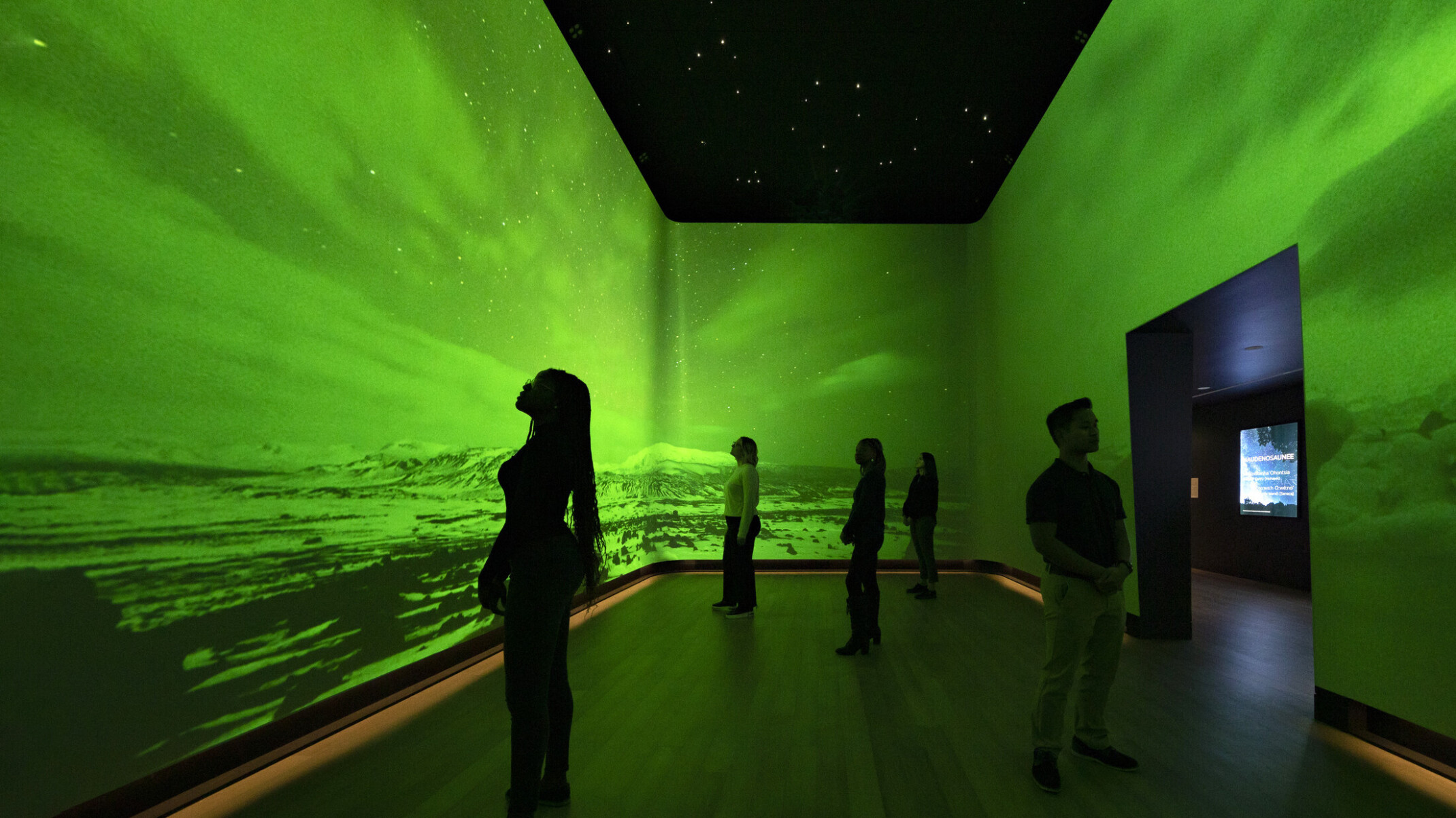 An immersive art exhibit at the Heard Museum with screens on all visible walls of the room showing the Northern Lights among snow-capped hills colorized with bright green