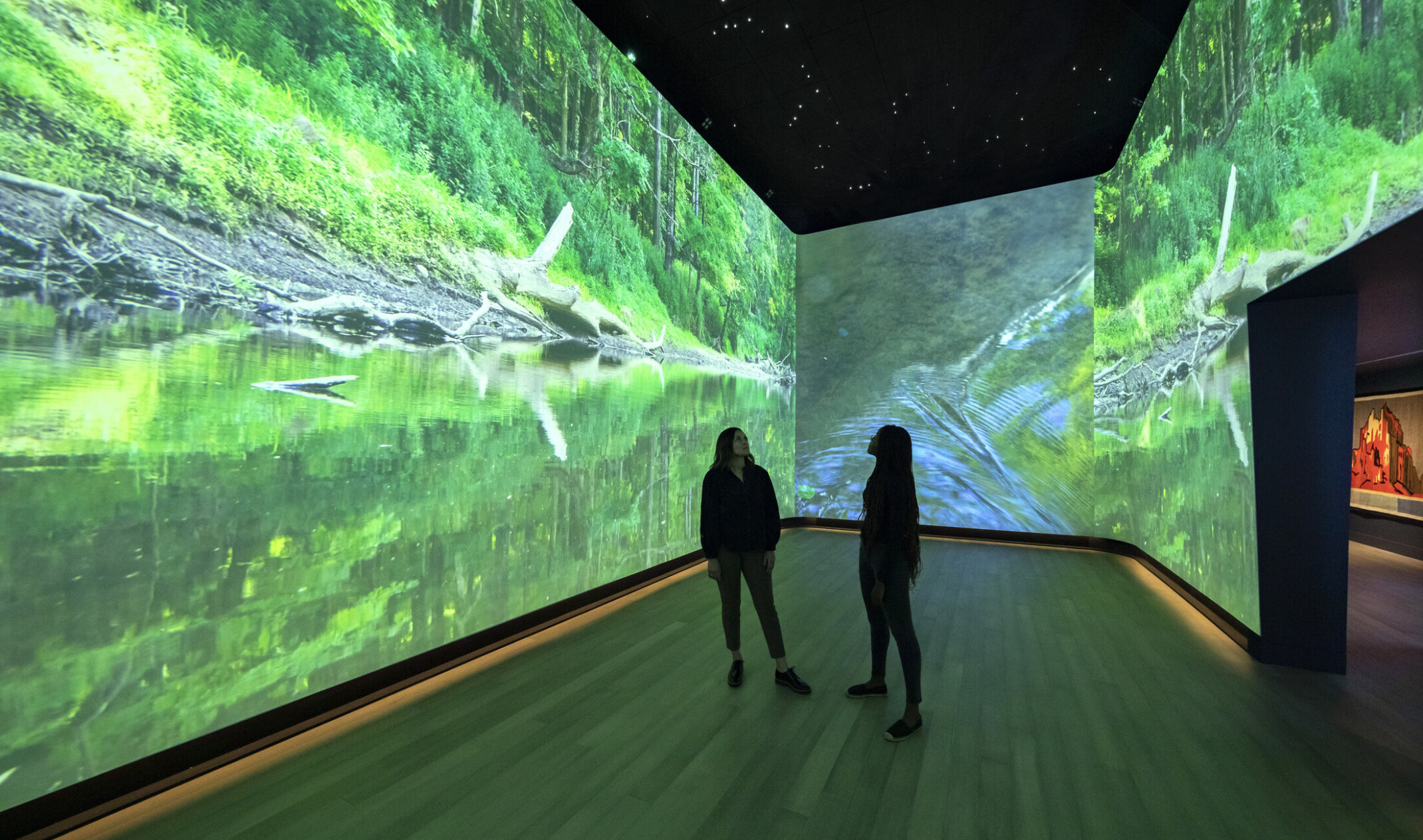 An immersive art exhibit at the Heard Museum with screens on all visible walls of the room showing a stream surrounded by greenery, interactive museum