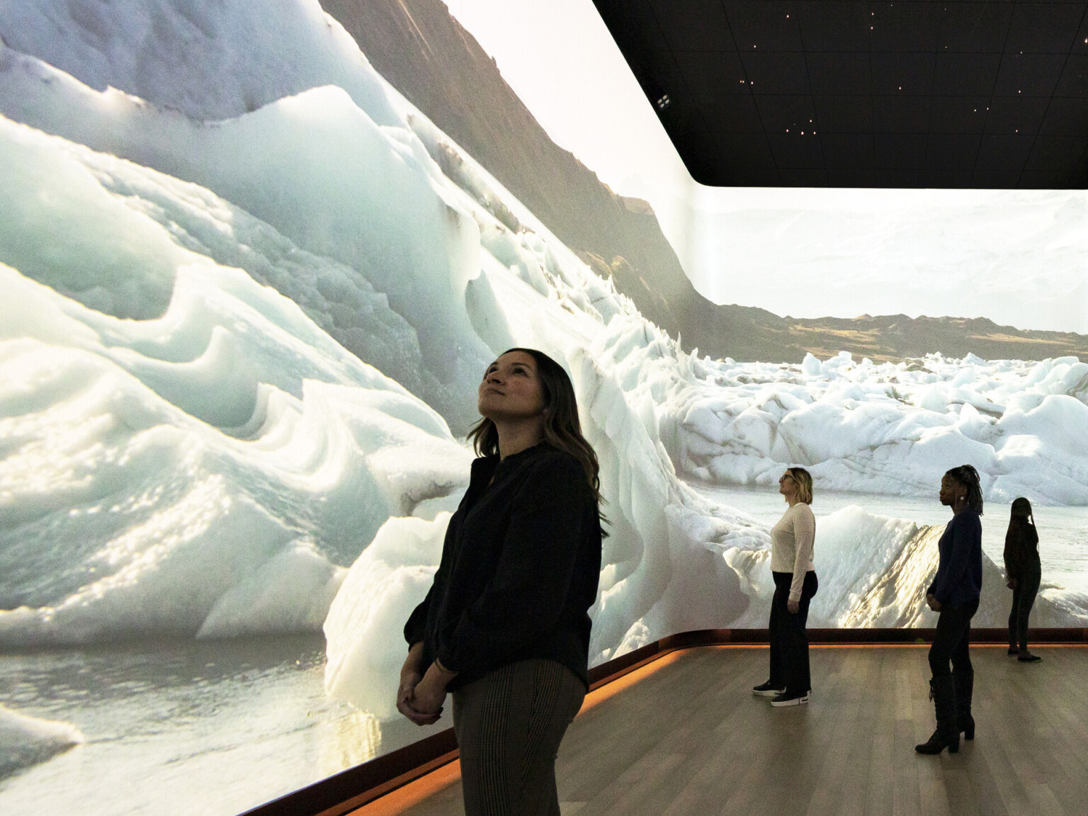 Woman stands in front of immersive 360 degree video art display of snow dunes and ice, interactive museum