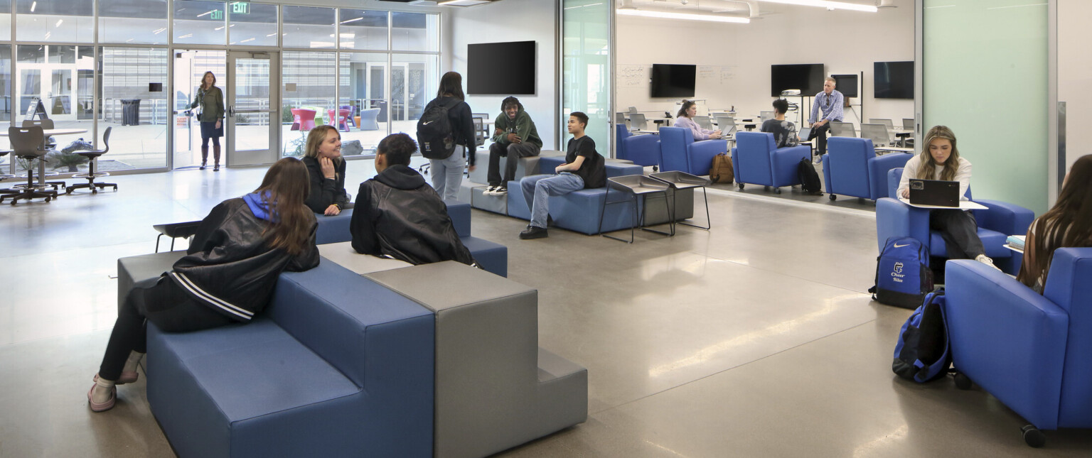 Classroom with large glass doors open to a spacious coworking area filled with students talking and working on comfy couches.