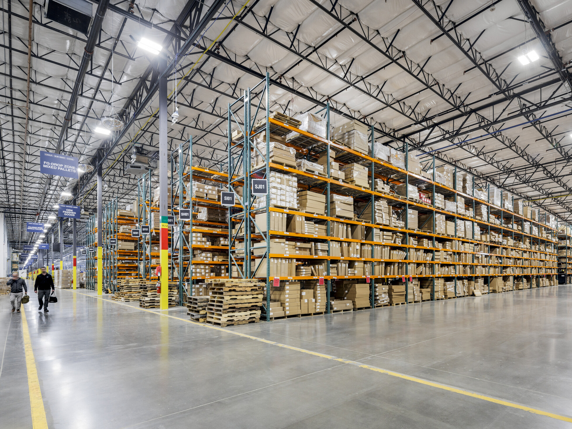Sub-Zero Group warehouse storage area; tall shelving units stacked with boxed inventory adjacent to a wide corridor; two workers walking through the space
