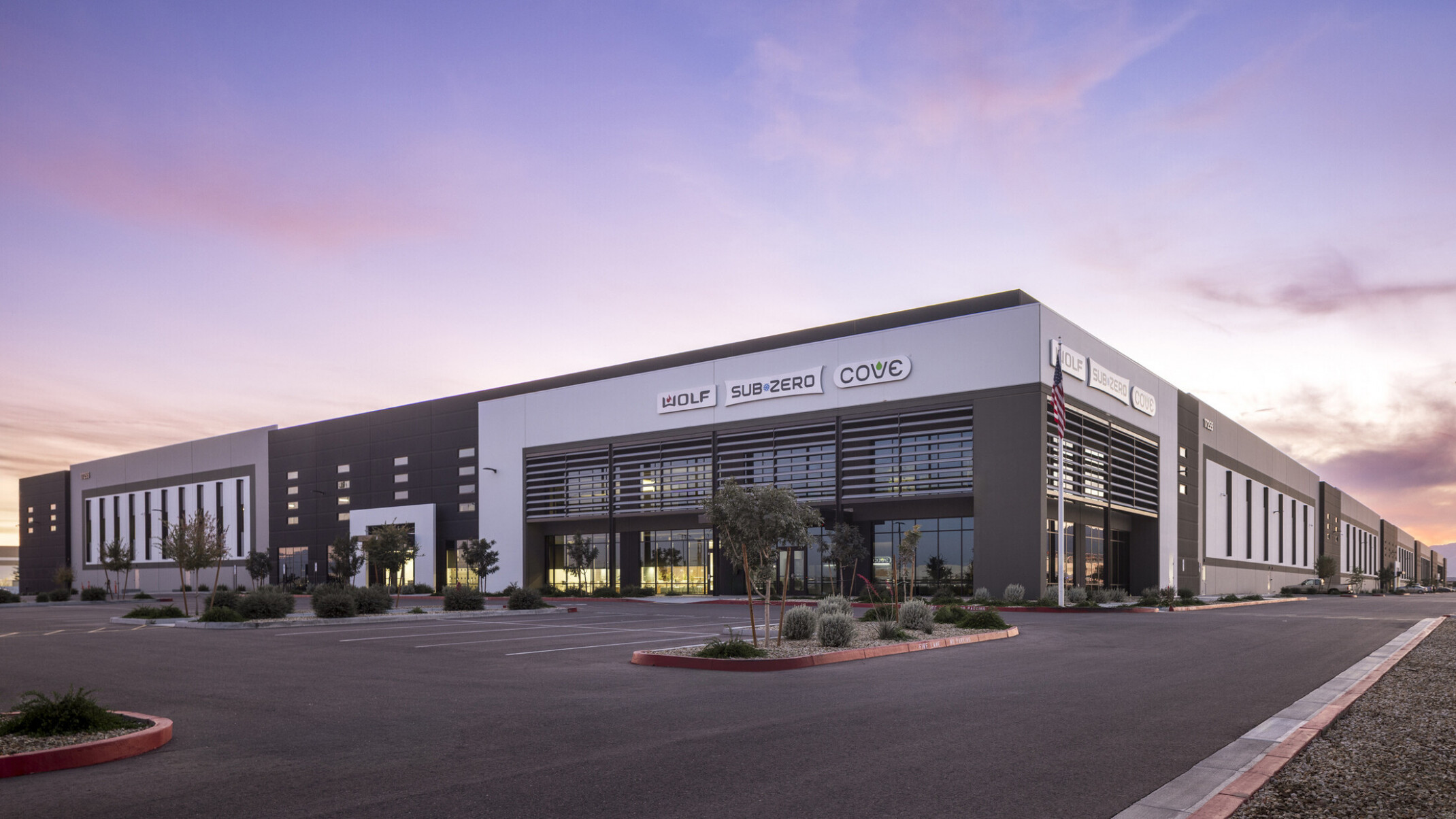 Sub-Zero Group facility exterior at dusk; white and slate tilt-up warehouse with large vertical louver fins framing the main entrance; Sub-Zero, Wolf, and Cove brand signage displayed on building facade; parking lot in foreground