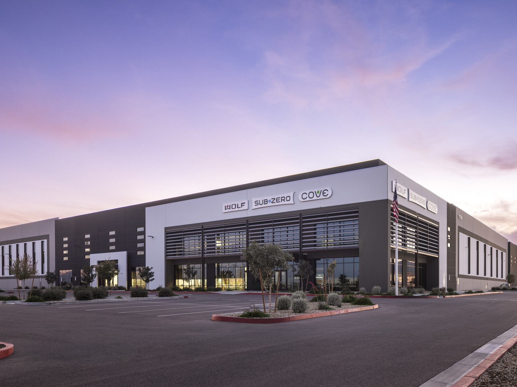 Sub-Zero Group facility exterior at dusk; white and slate tilt-up warehouse with large vertical louver fins framing the main entrance; Sub-Zero, Wolf, and Cove brand signage displayed on building facade; parking lot in foreground