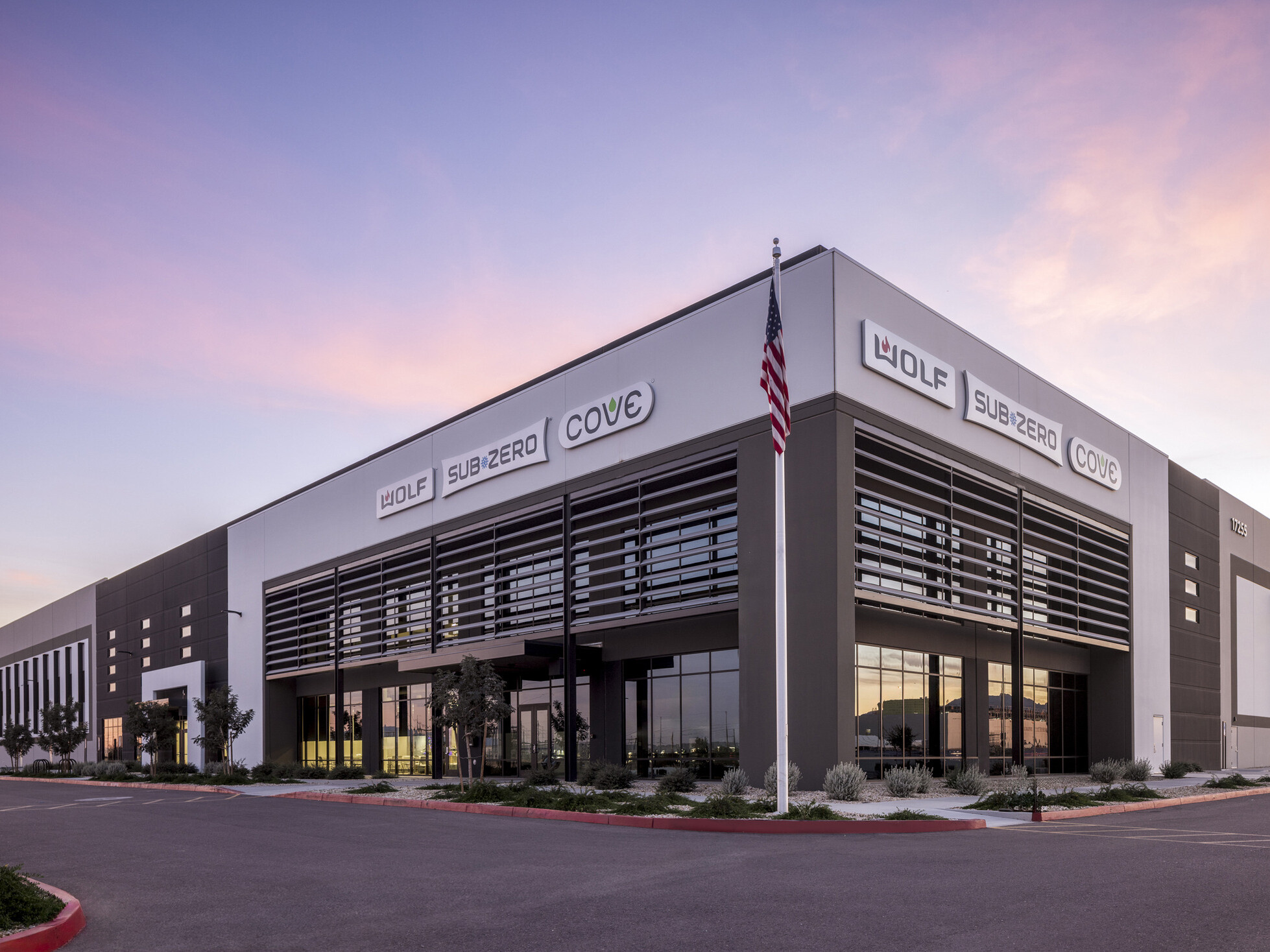 Sub-Zero Group facility exterior at dusk; white and slate tilt-up warehouse with large vertical louver fins framing the main entrance; Sub-Zero, Wolf, and Cove brand signage displayed on building facade; parking lot in foreground