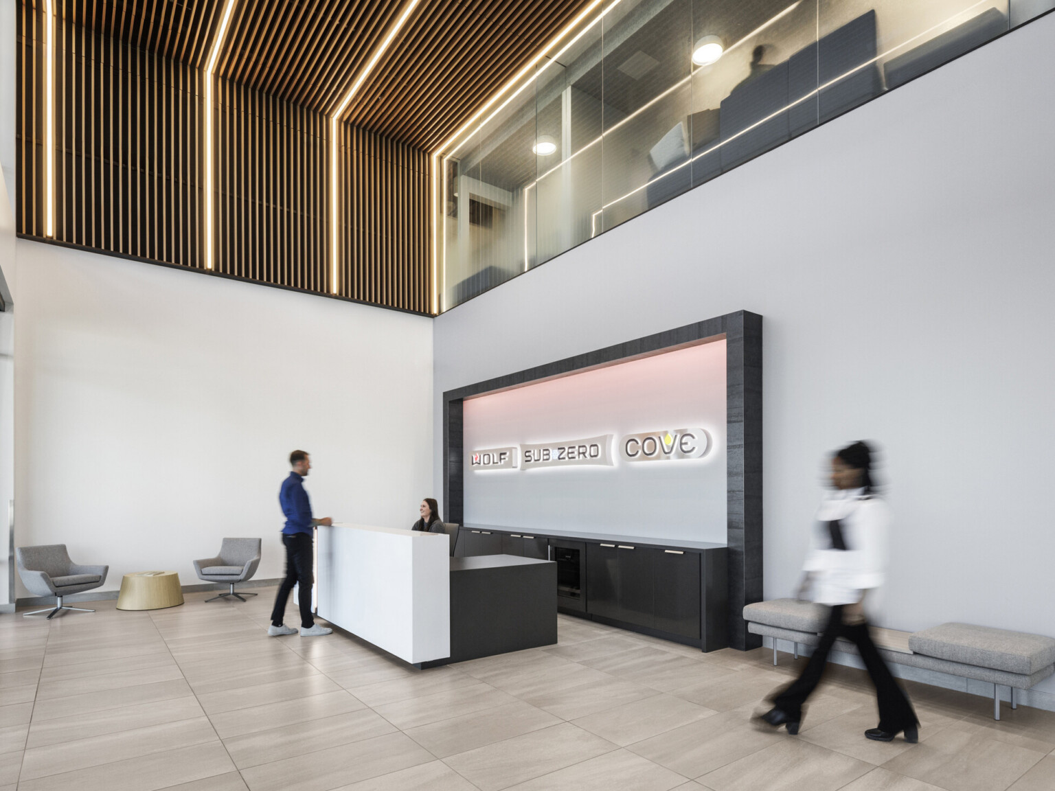 Sub-Zero Group reception area; black and white reception desk with framed accent wall displaying Sub-Zero, Wolf, and Cove logos washed in light; two-story lobby with wood slat wall-to-ceiling system wrapping