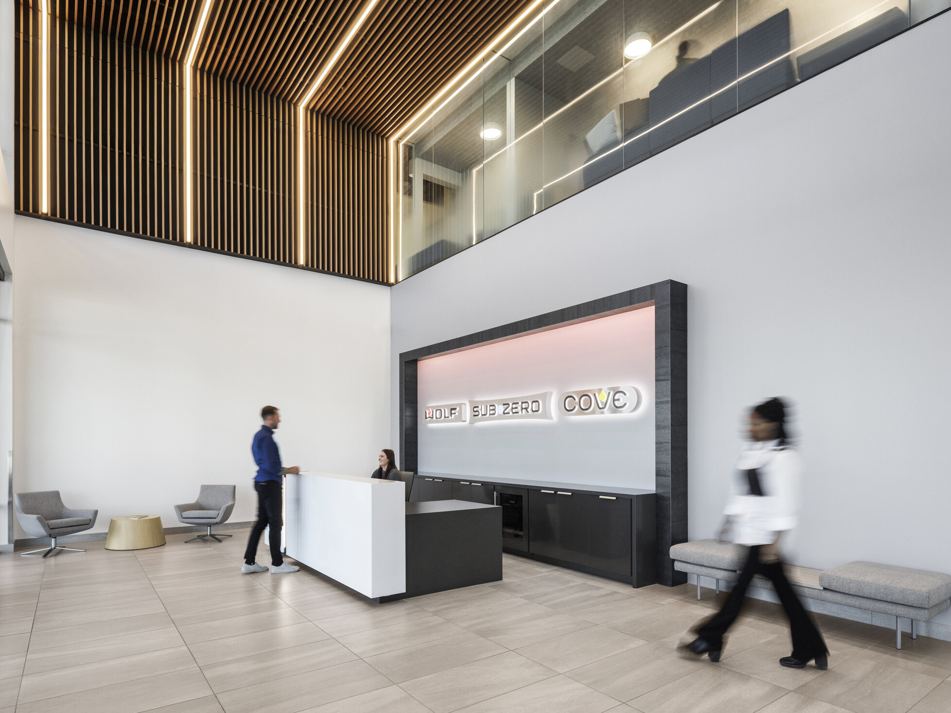 Sub-Zero Group reception area; black and white reception desk with framed accent wall displaying Sub-Zero, Wolf, and Cove logos washed in light; two-story lobby with wood slat wall-to-ceiling system wrapping